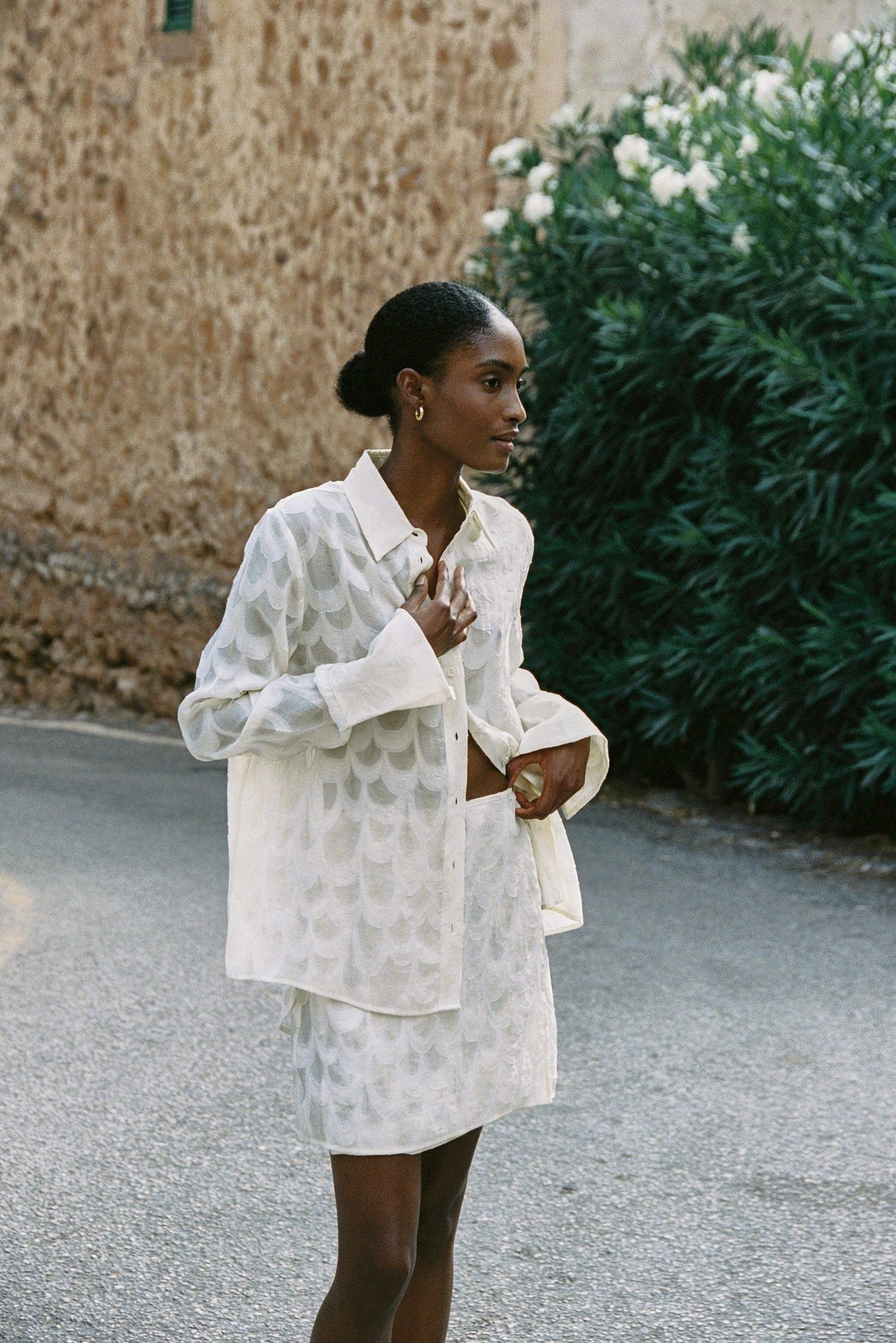 Model wearing the AUDĒRE. Rae semi-sheer shirt in Ivory, standing beside a flowering wall.