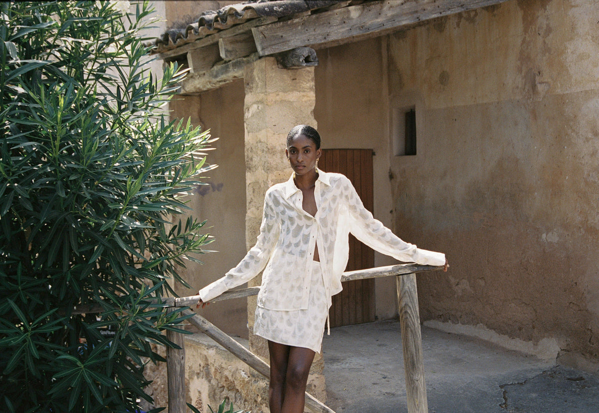 Model wearing the AUDĒRE. Rae semi-sheer shirt and wrap mini skirt in Ivory beside a stone stairway.