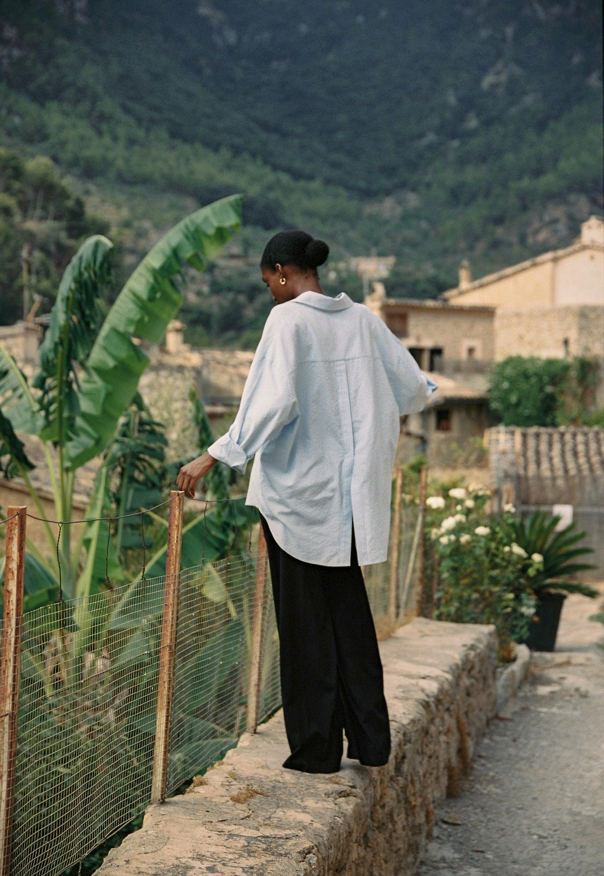 Model wearing the AUDĒRE. Alba oversized striped shirt in Ciel blue, back view standing beside a stone wall.
