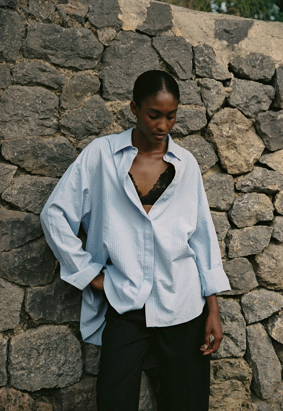 Model wearing the AUDĒRE. Alba oversized striped shirt in Ciel blue, close-up beside a stone wall.