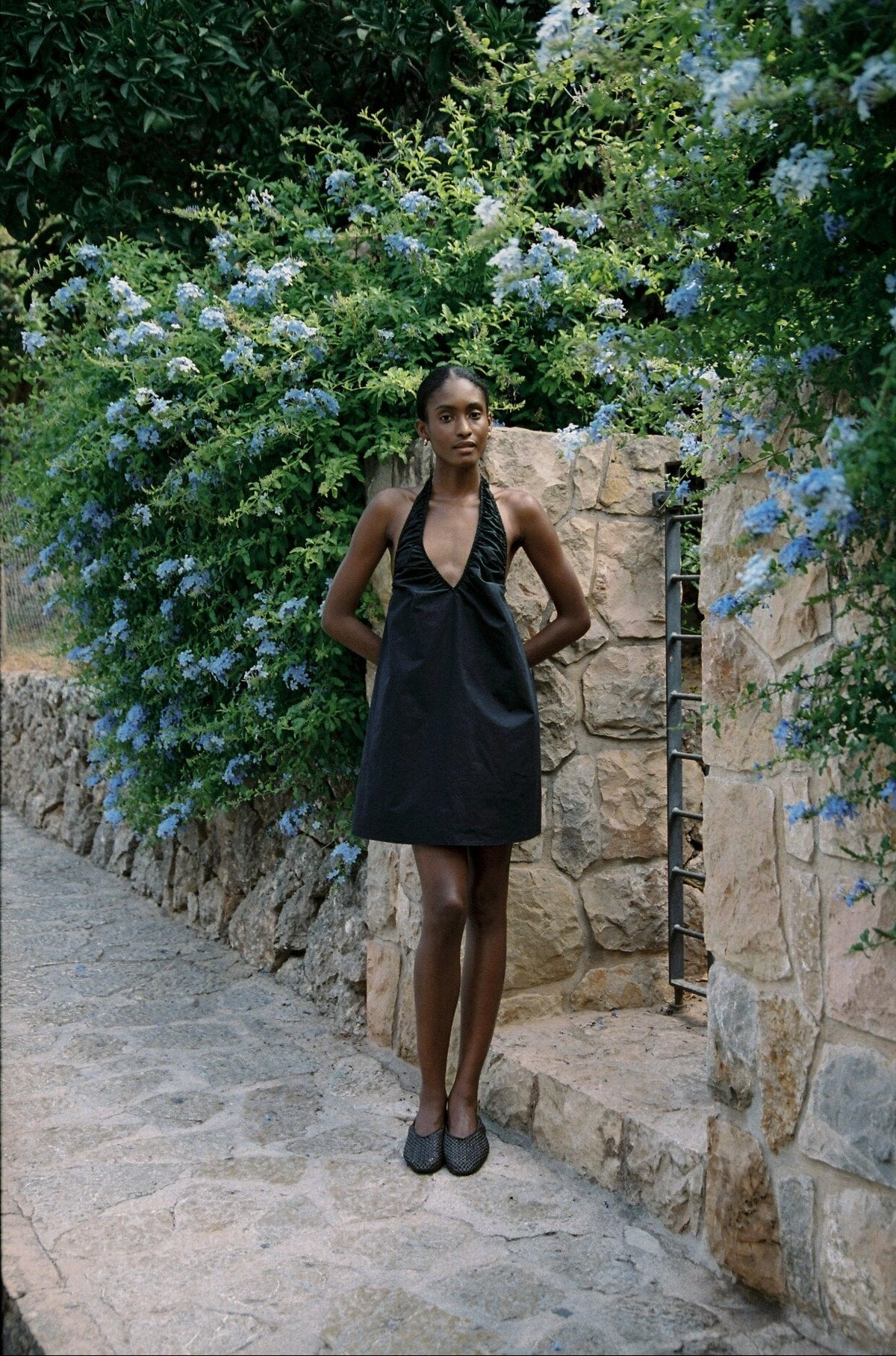 Model wearing the AUDĒRE. Azura cotton halter mini dress in Black, full-length view in a stone lane.