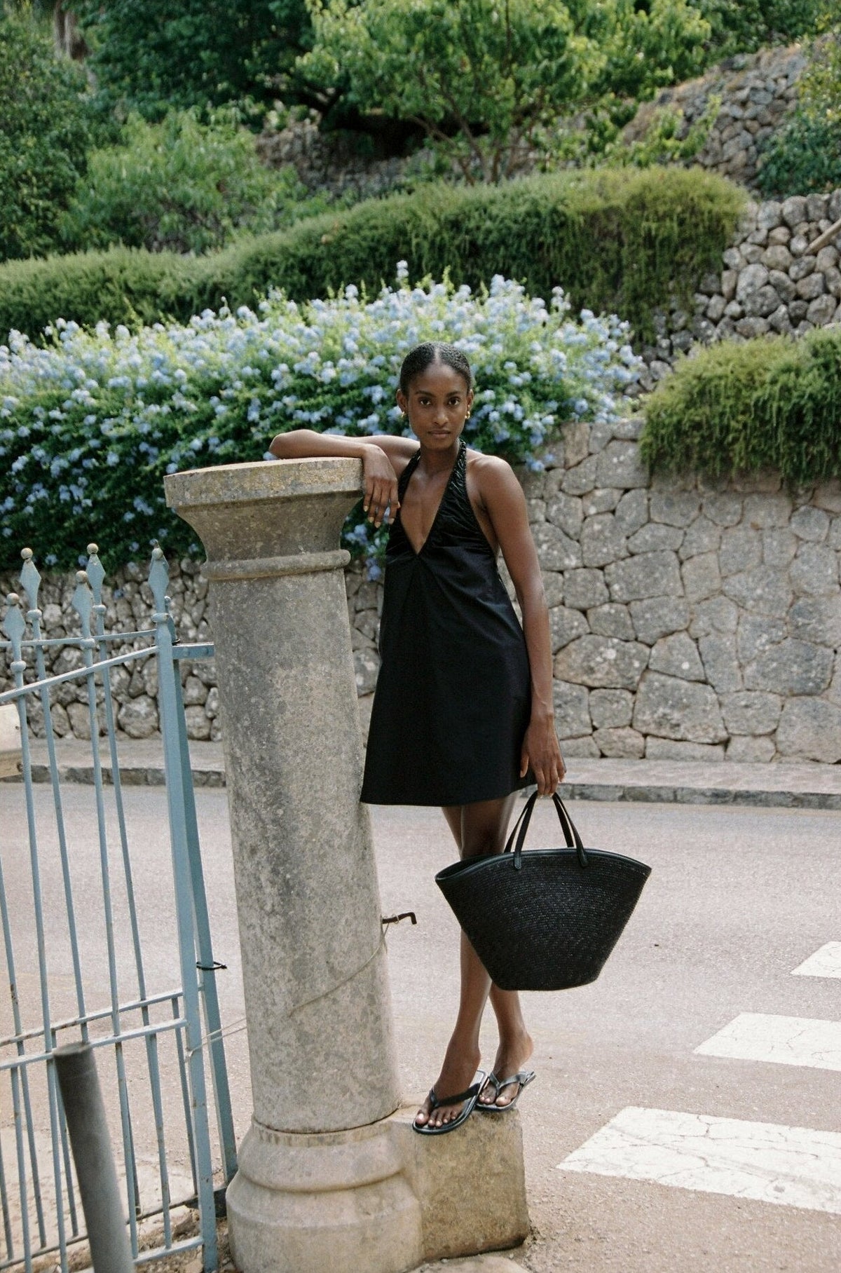 Model wearing the AUDĒRE. Azura cotton halter mini dress in Black, full-length side view with a black tote.