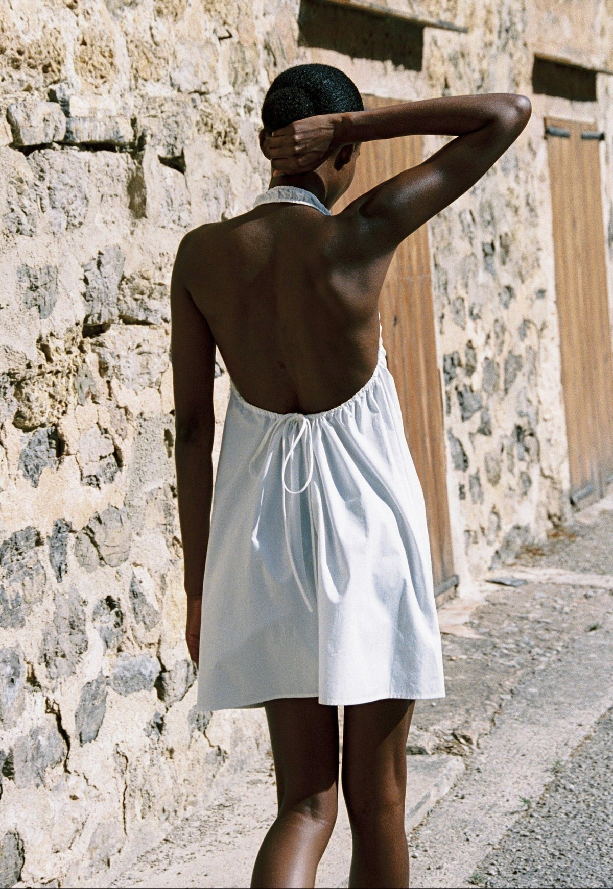 Model wearing the AUDĒRE. Azura cotton halter mini dress in White, back view showing the open-back halter cut.