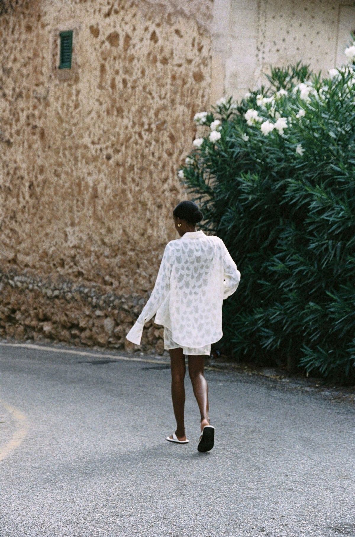 Model wearing the AUDĒRE. Rae semi-sheer shirt in Ivory, back view while walking along a stone lane.