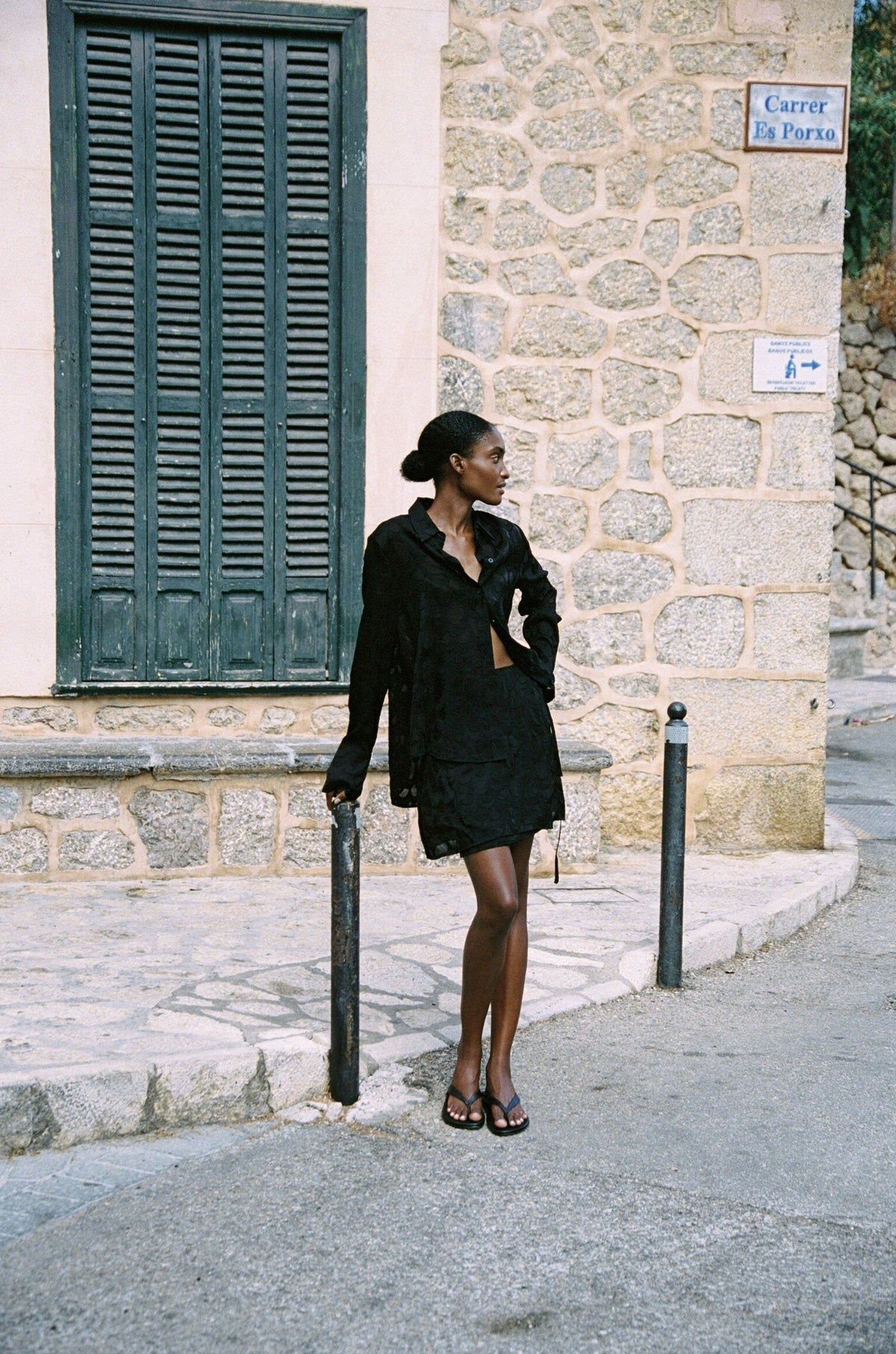 Model wearing the AUDĒRE. Rae semi-sheer shirt in Black, full-length side view beside green shutters.