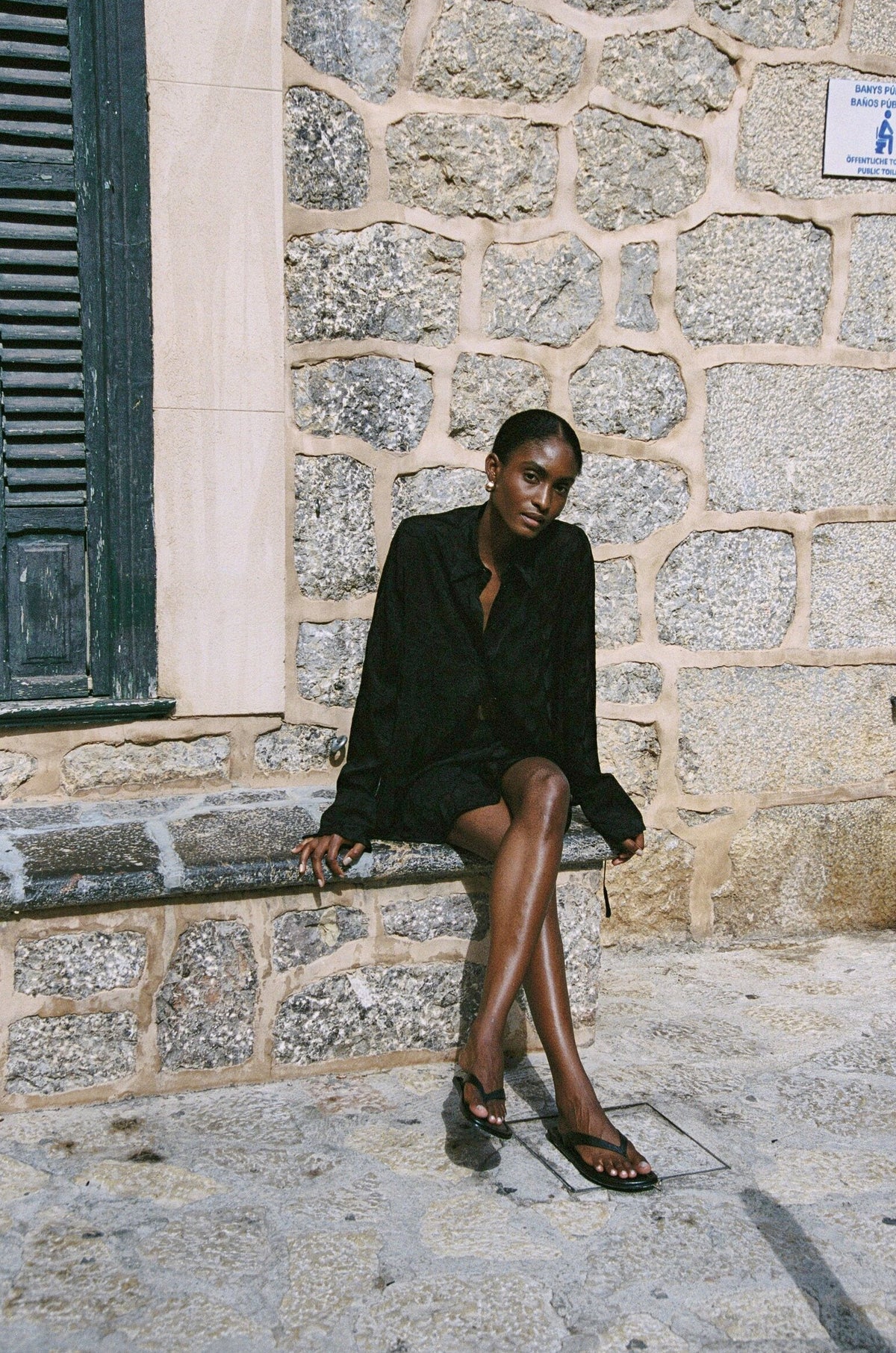 Model wearing the AUDĒRE. Rae semi-sheer shirt in Black, seated beside green shutters.