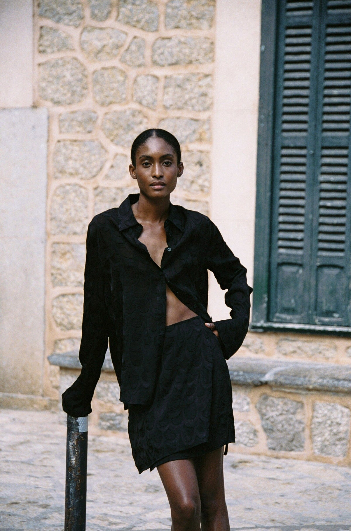 Model wearing the AUDĒRE. Rae wrap mini skirt in Black, front view with the matching shirt in a stone lane.