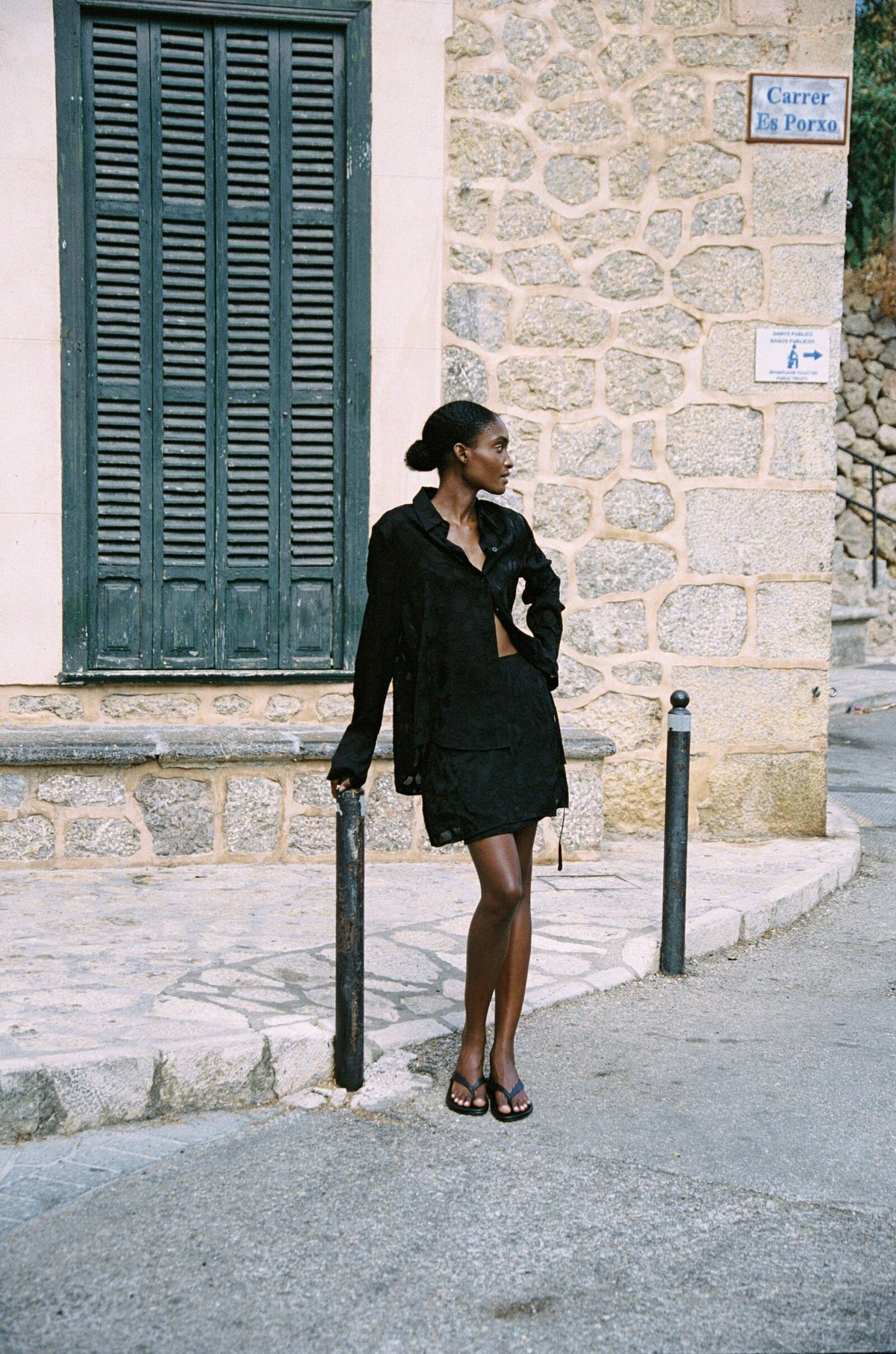 Model wearing the AUDĒRE. Rae wrap mini skirt in Black, full-length view beside green shutters.