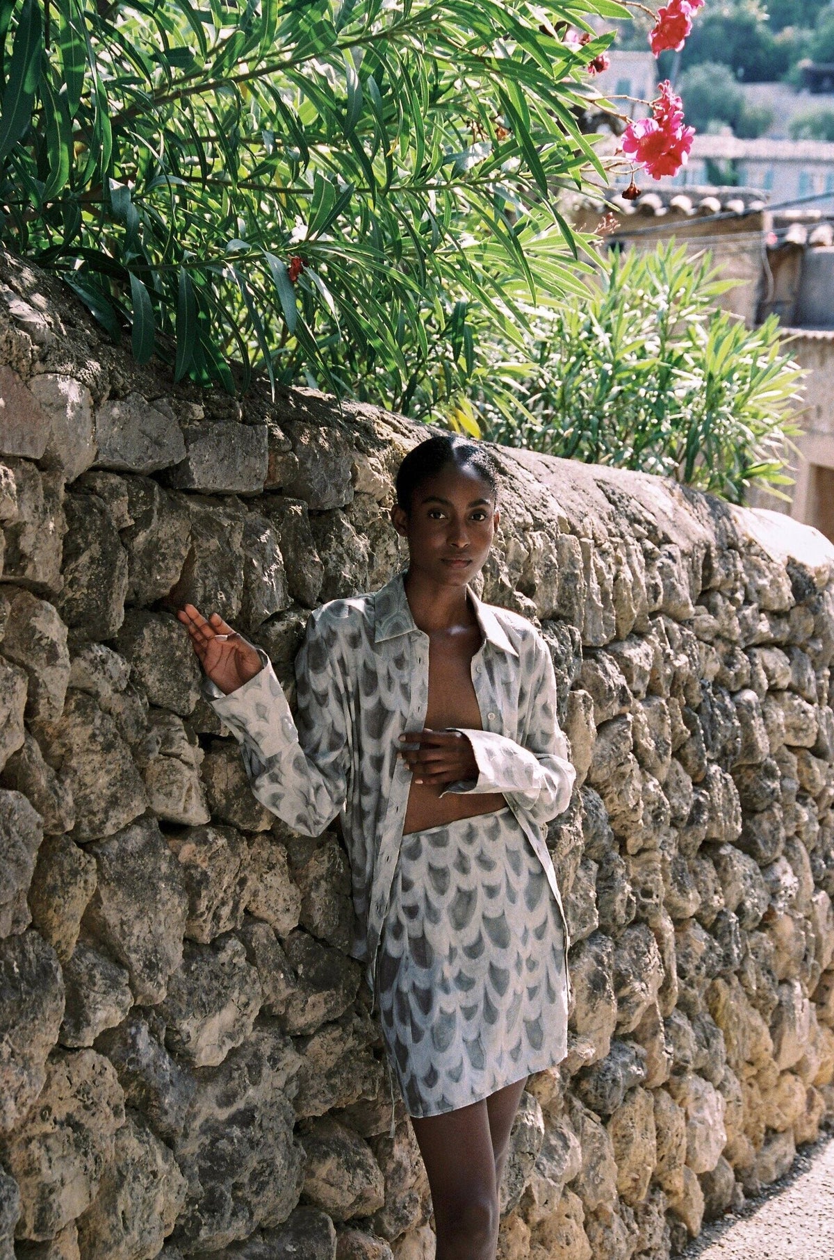 Model wearing the AUDĒRE. Rae wrap mini skirt in Dove, standing beside a stone wall with the matching shirt.
