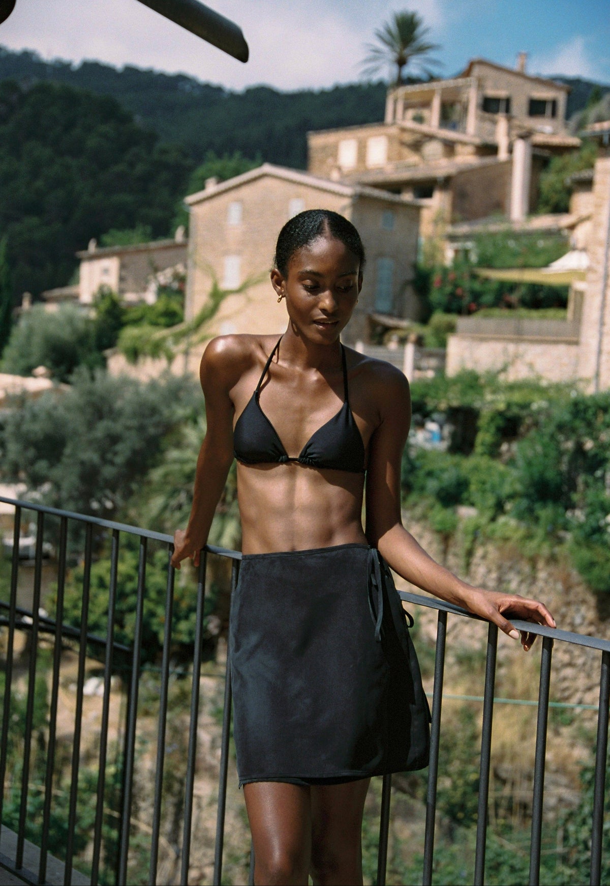 Model wearing the AUDĒRE. Vero cupro-blend wrap mini skirt in Black, standing beside a railing with a black bralette.