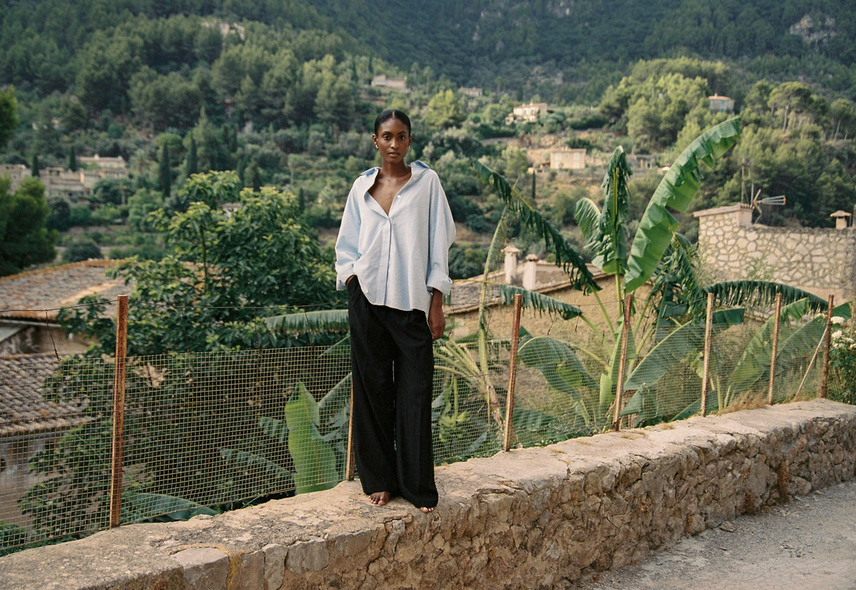 Model wearing the AUDĒRE. Alba oversized striped shirt in Ciel blue with black trousers overlooking a village.