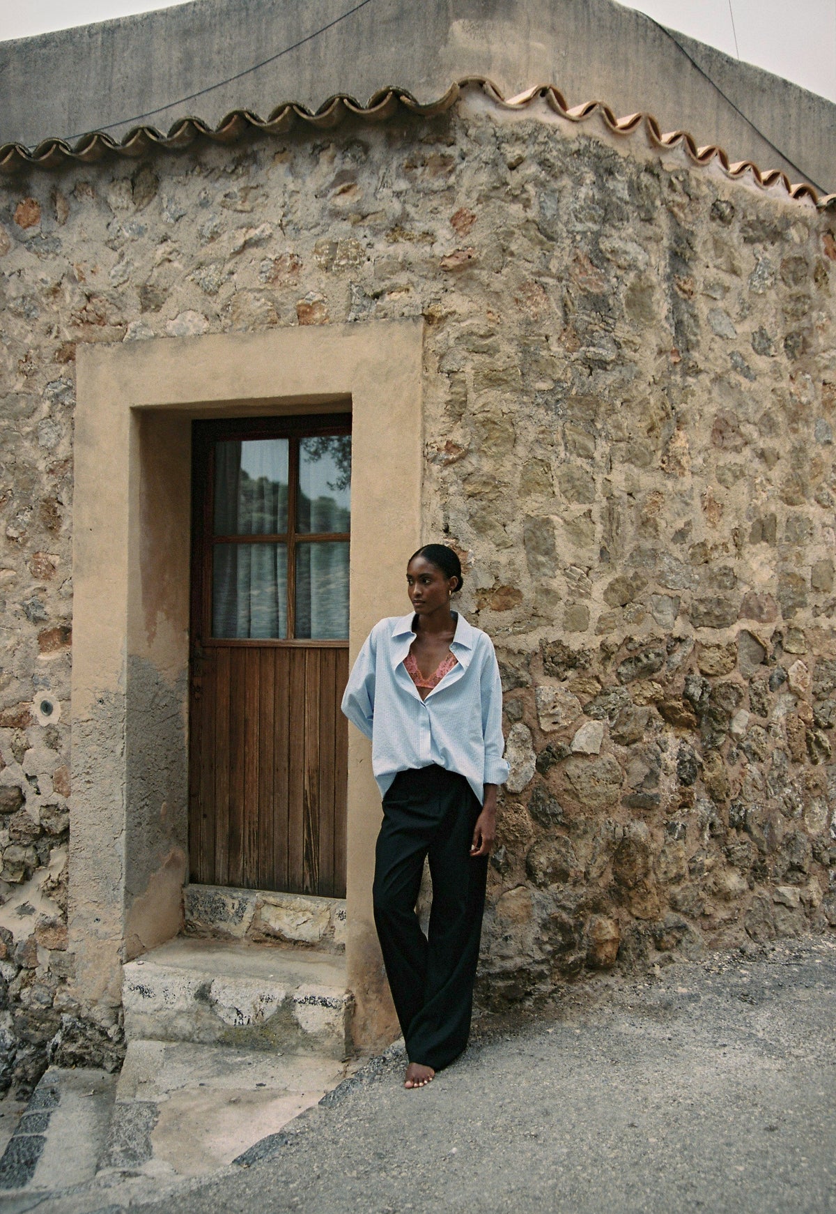 Model wearing the AUDĒRE. Alba oversized striped shirt in Ciel blue, full-length view beside a stone building.