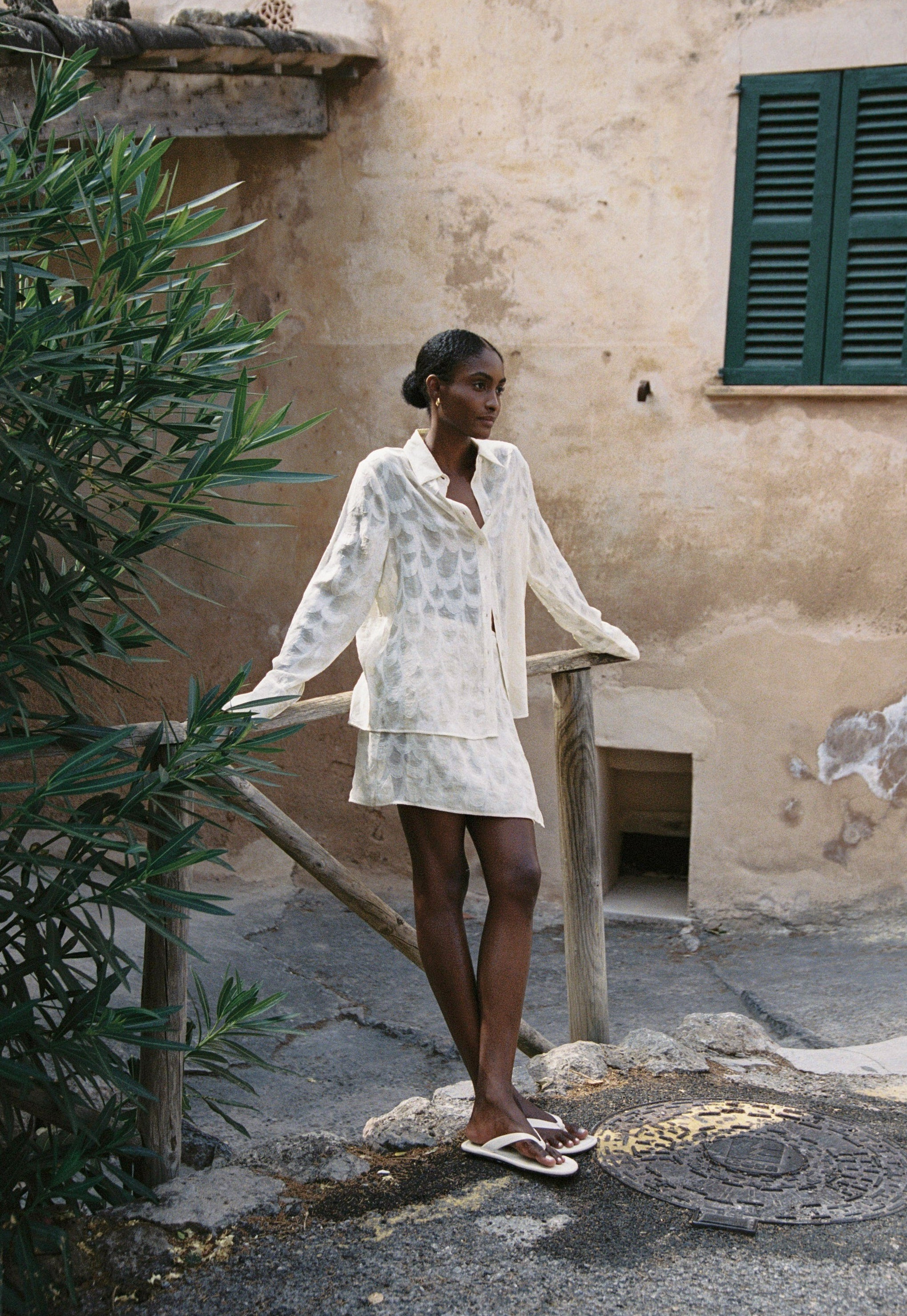 Model wearing the AUDĒRE. Rae semi-sheer shirt and wrap mini skirt in Ivory, full-length view beside a stone stairway.