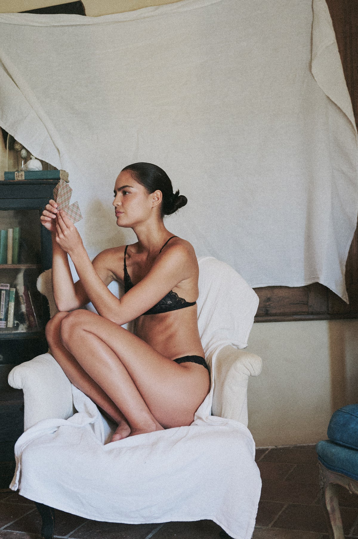 Model wearing the AUDĒRE. Noemi black lace bralette and matching bottoms, seated in a white armchair.