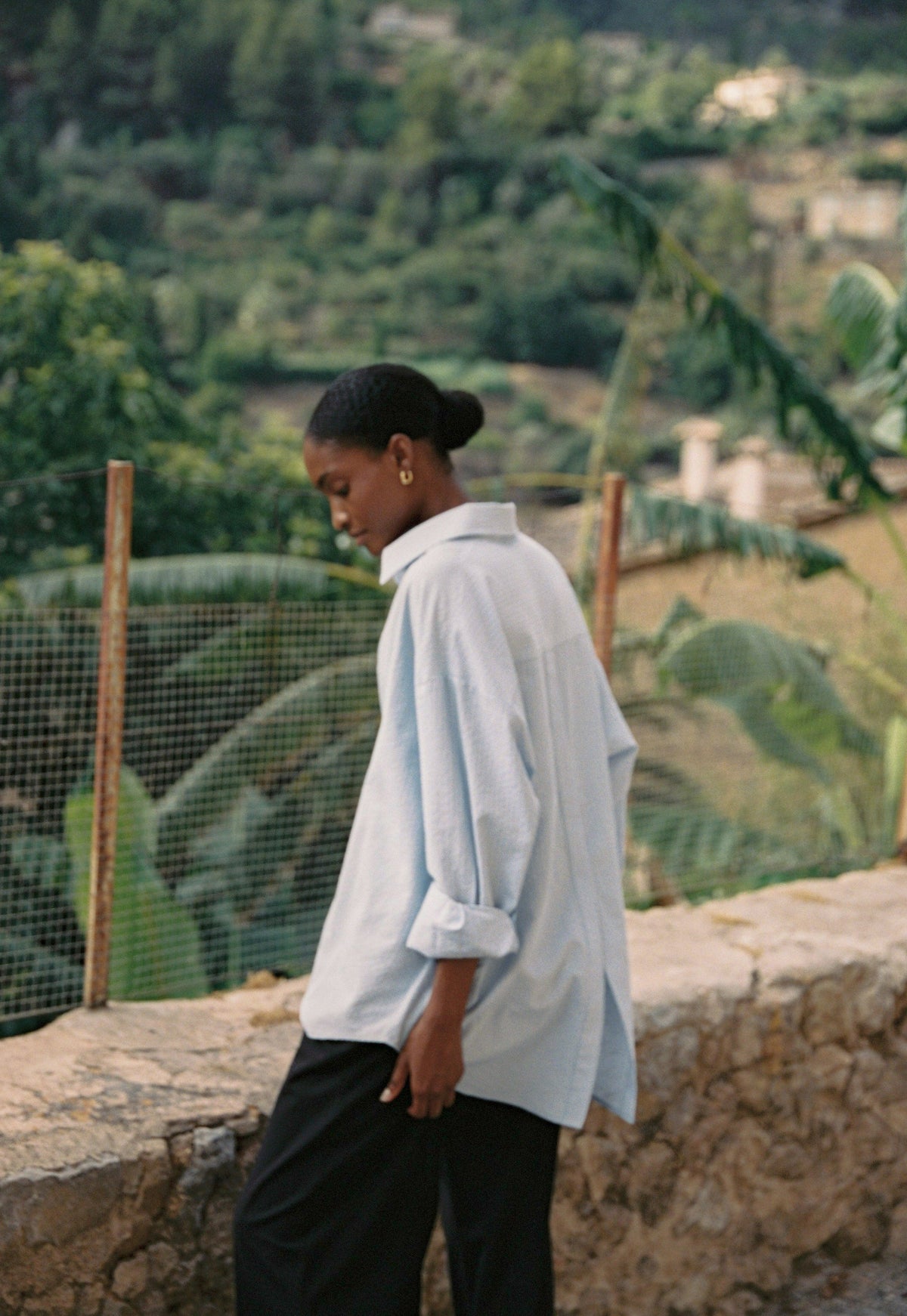 Model wearing the AUDĒRE. Alba oversized striped shirt in Ciel blue, back view overlooking a village.