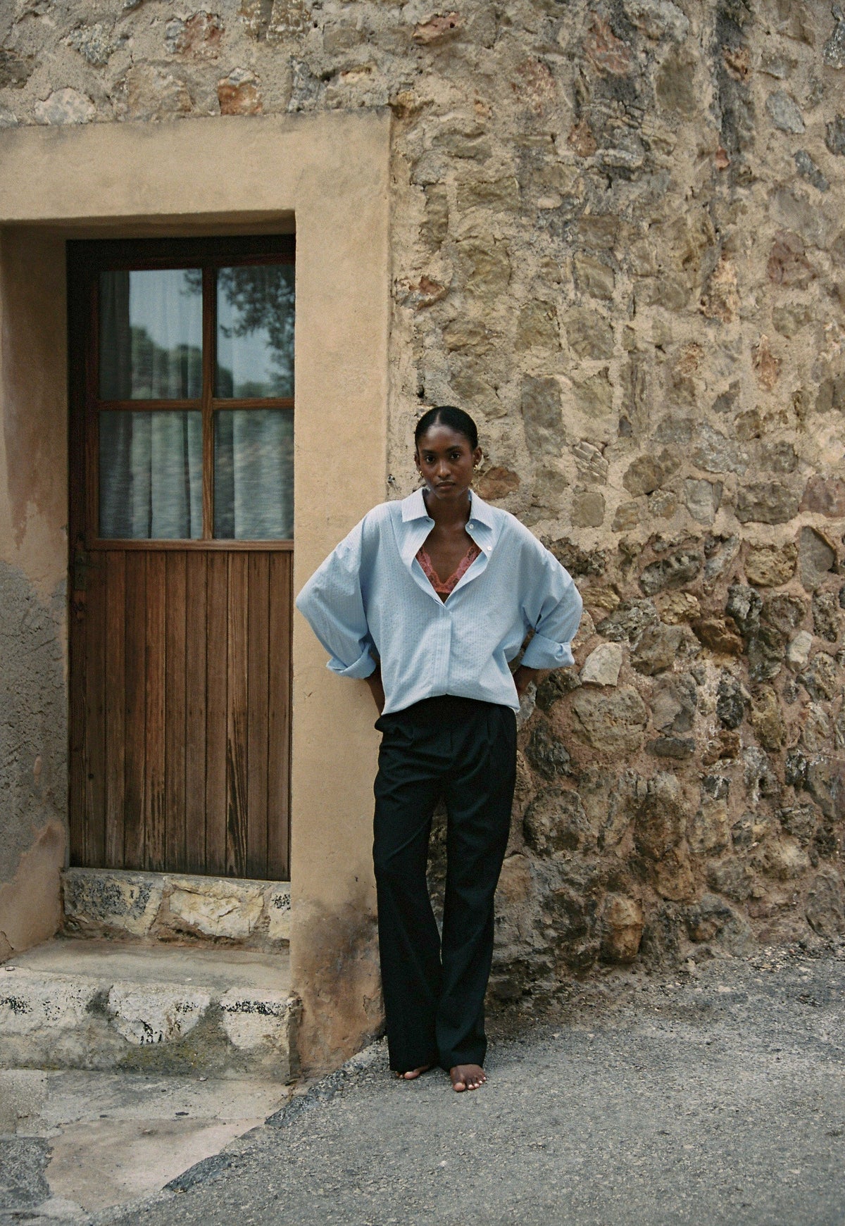 Model wearing the AUDĒRE. Alba oversized striped shirt in Ciel blue, standing beside a stone doorway.
