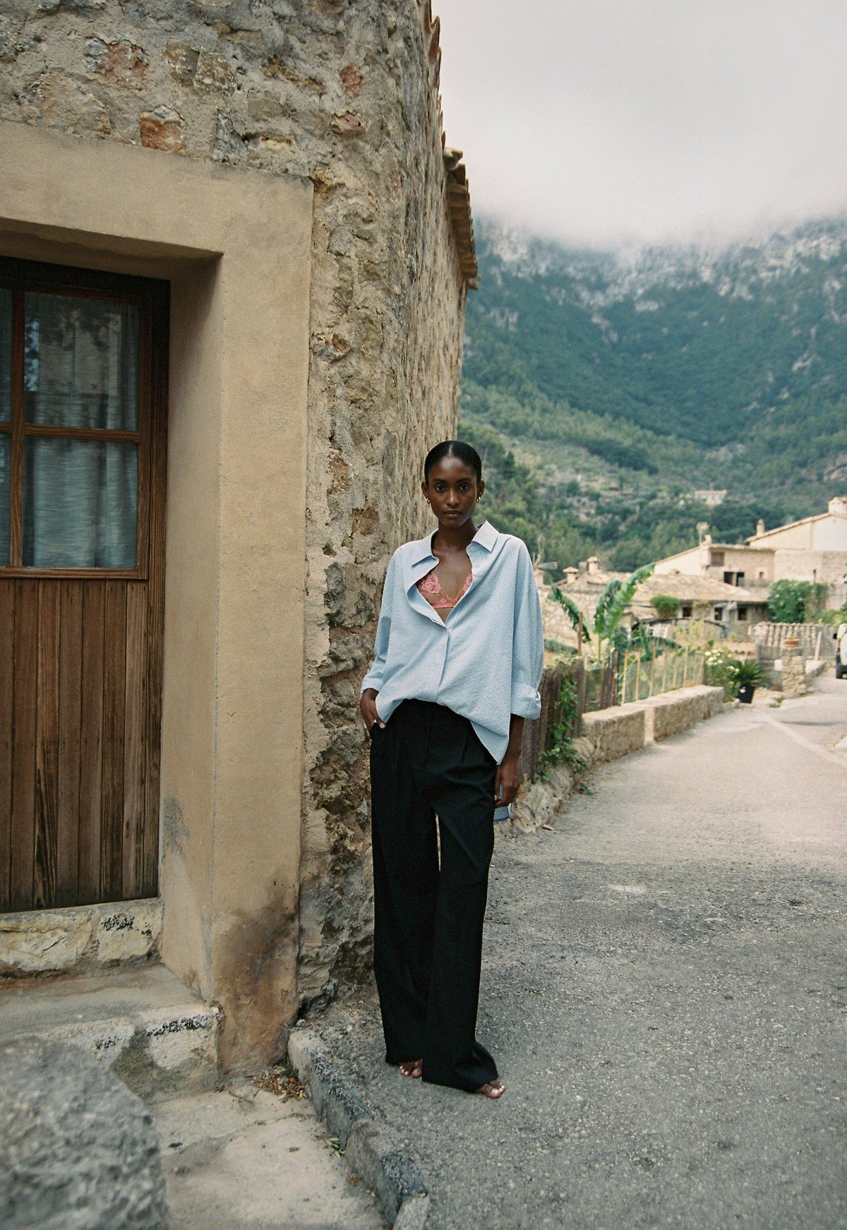 Model wearing the AUDĒRE. Alba oversized striped shirt in Ciel blue, standing beside a stone wall overlooking a village.