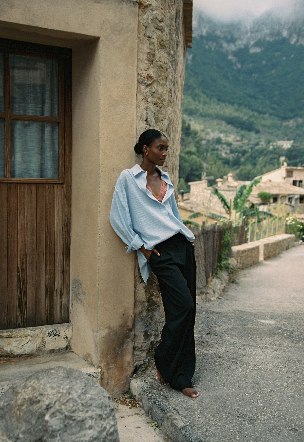 Model wearing the AUDĒRE. Alba oversized striped shirt in Ciel blue, leaning against a stone doorway.