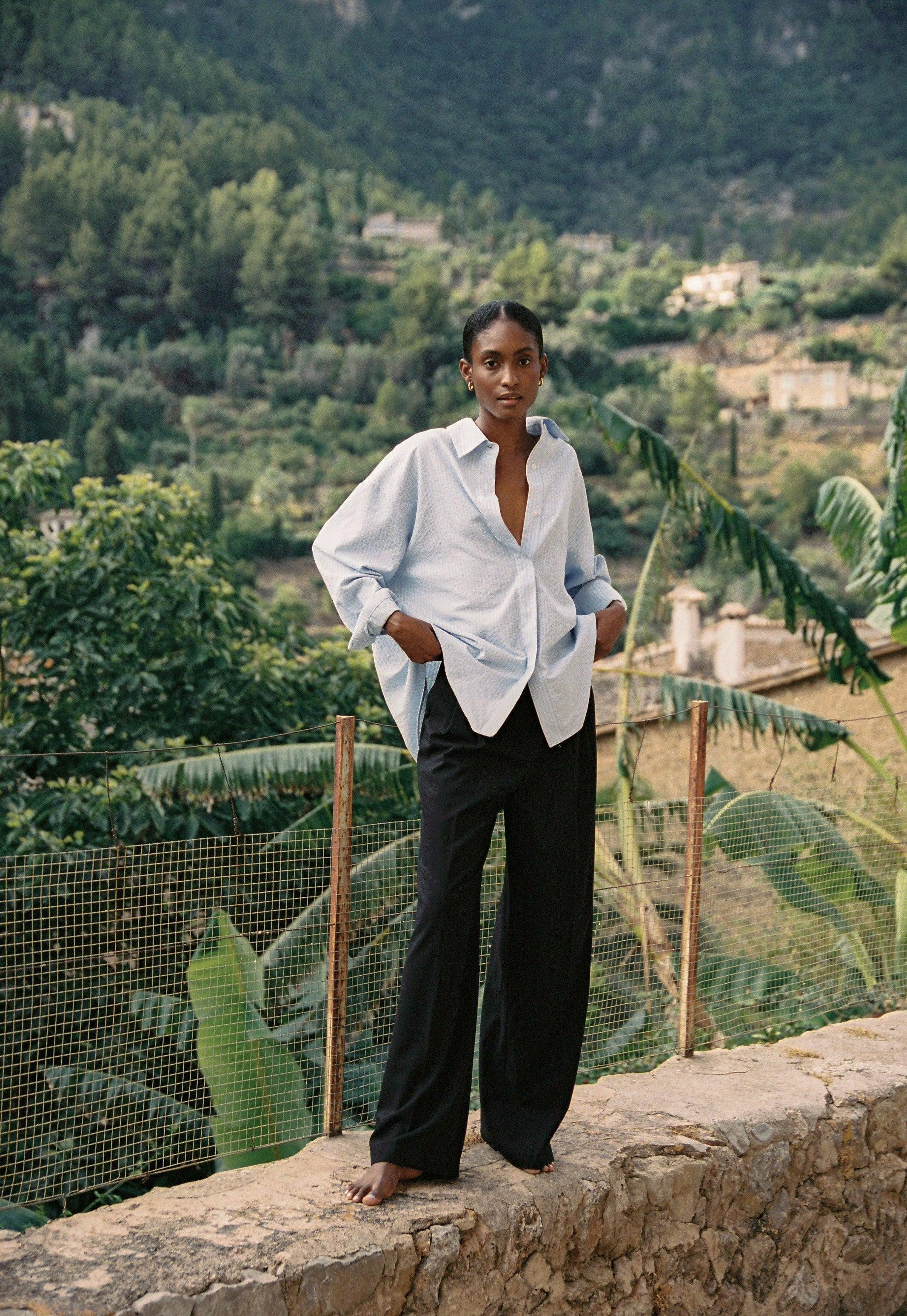 Model wearing the AUDĒRE. Alba oversized striped shirt in Ciel blue, front view overlooking a village.