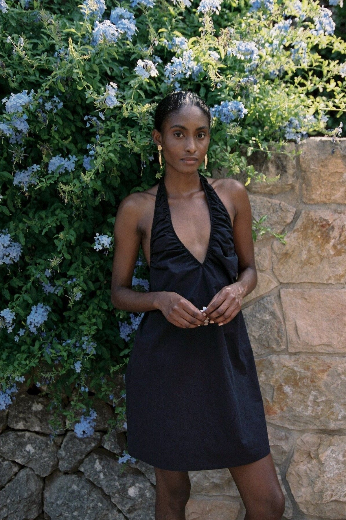 Model wearing the AUDĒRE. Azura cotton halter mini dress in Black, standing beside a flowering stone wall.