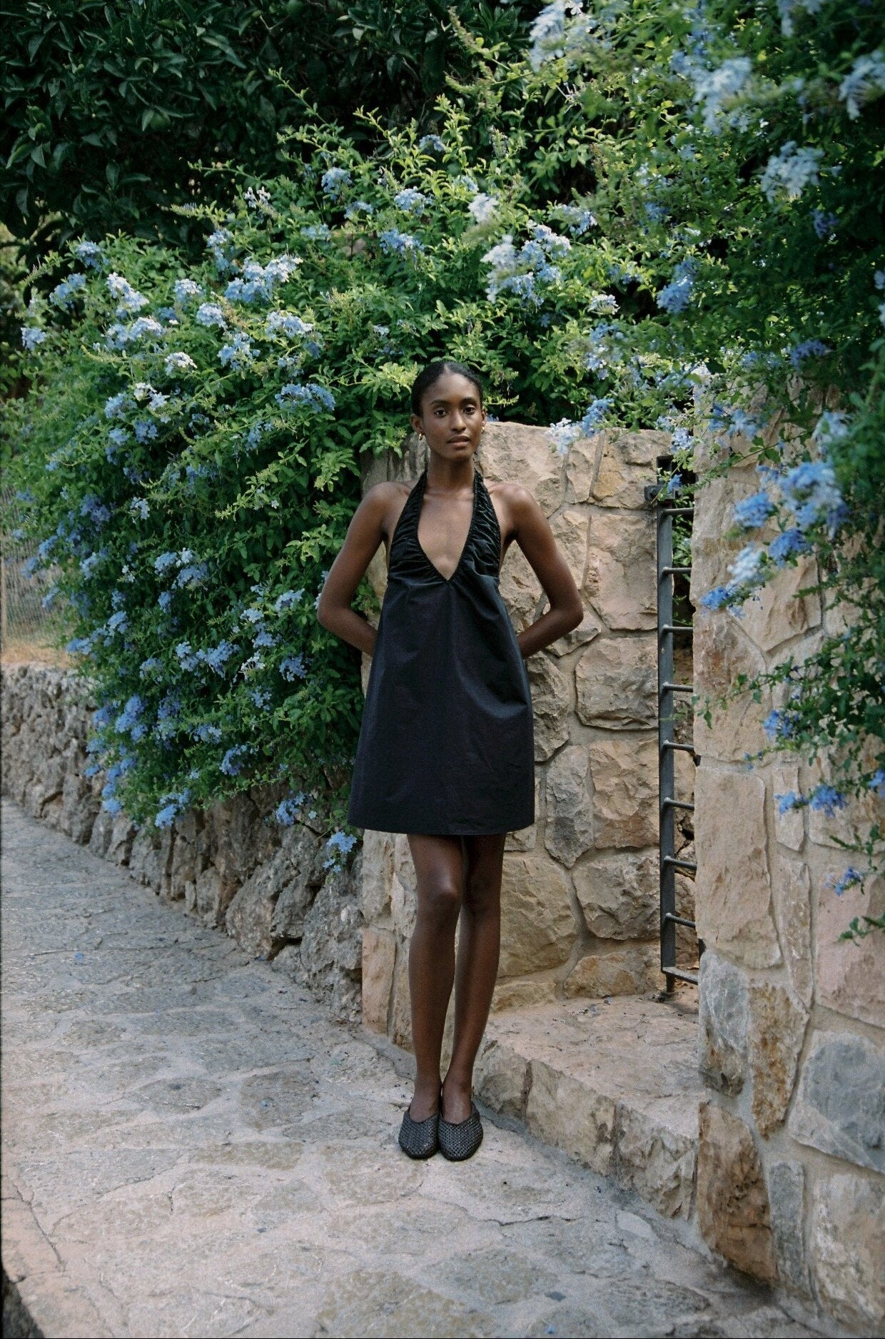Model wearing the AUDĒRE. Azura cotton halter mini dress in Black, full-length view in a stone lane.