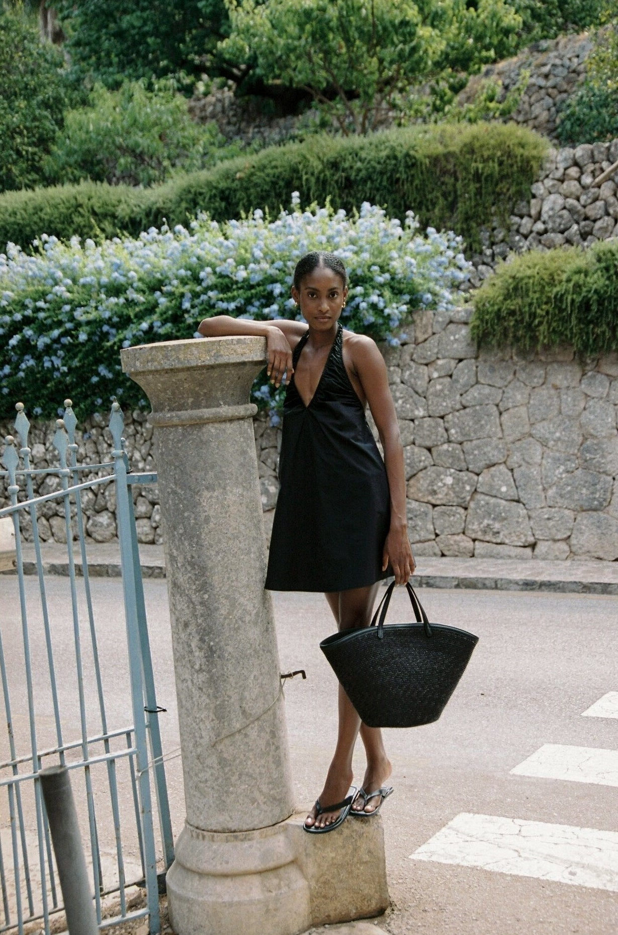Model wearing the AUDĒRE. Azura cotton halter mini dress in Black, full-length side view with a black tote.