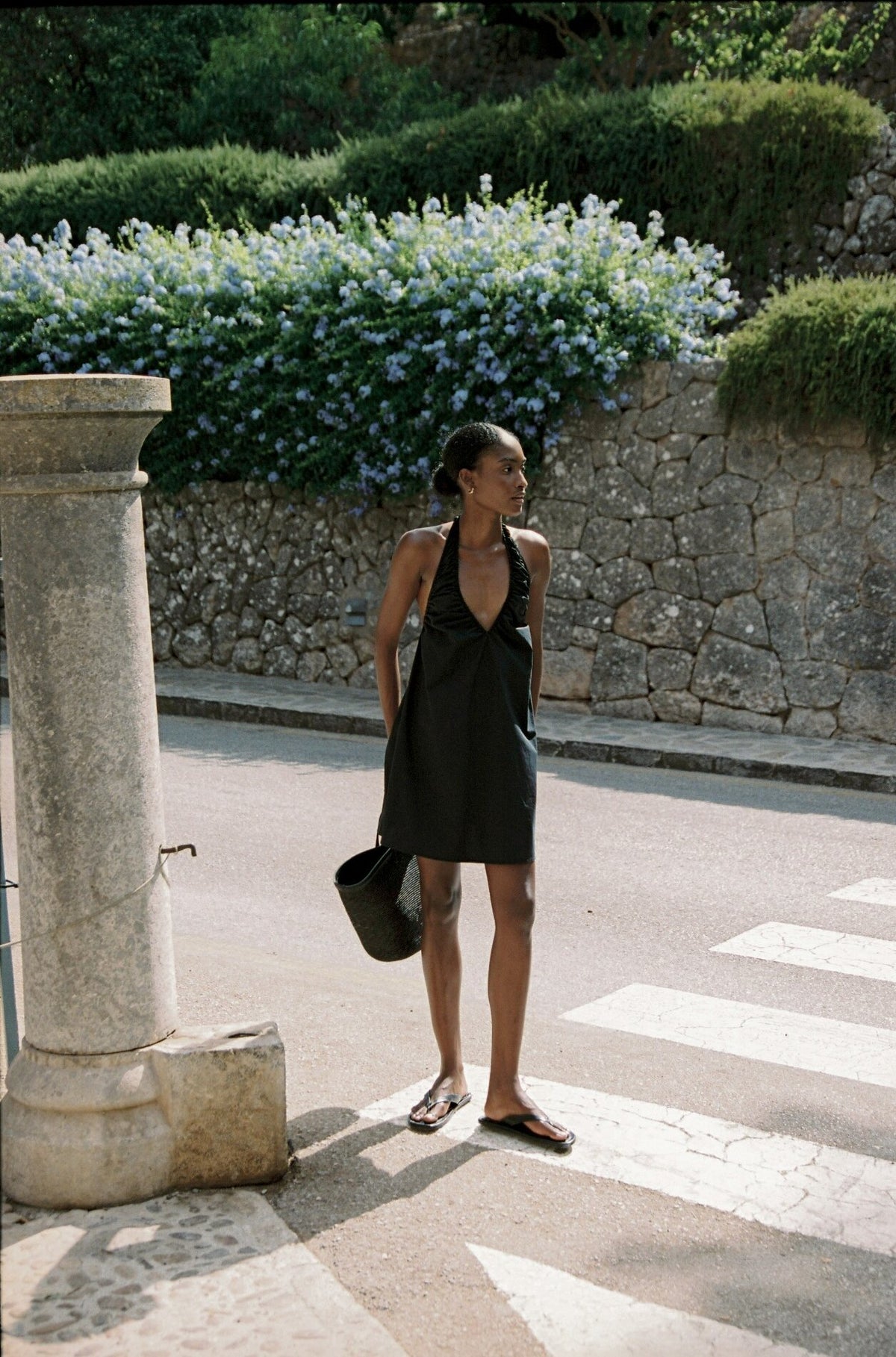 Model wearing the AUDĒRE. Azura cotton halter mini dress in Black, walking beside a stone wall with a black tote.