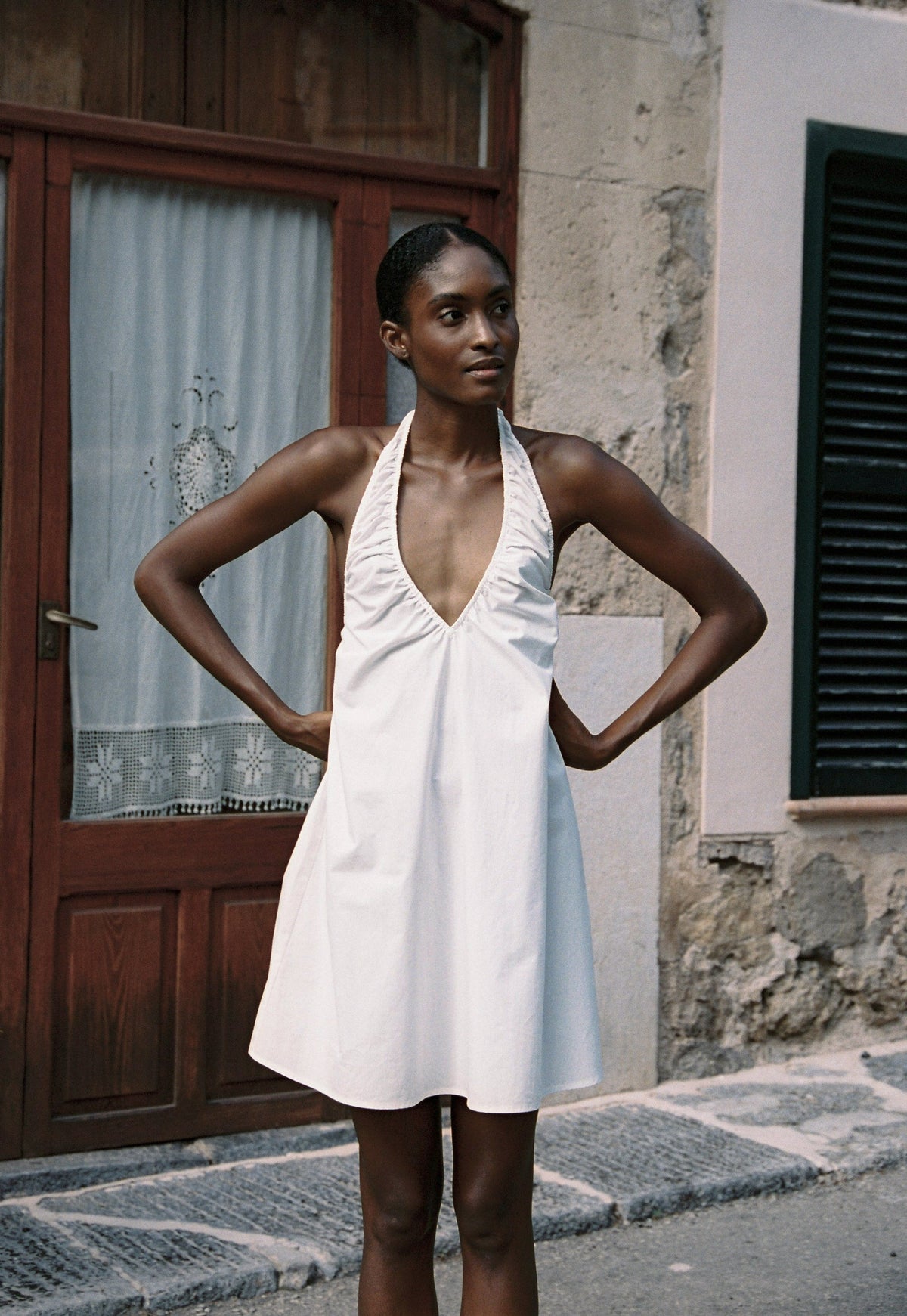 Model wearing the AUDĒRE. Azura cotton halter mini dress in White, front view with hands on hips.