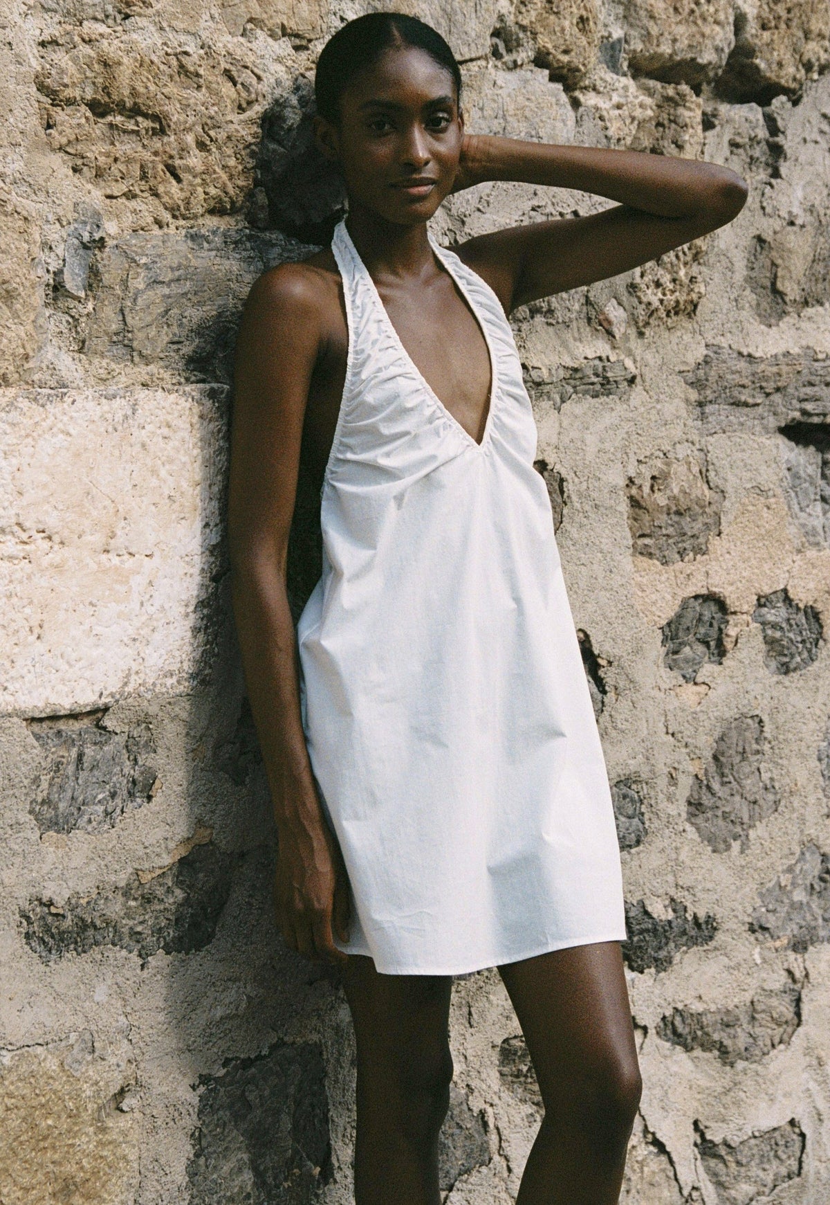 Model wearing the AUDĒRE. Azura cotton halter mini dress in White, standing against a stone wall.