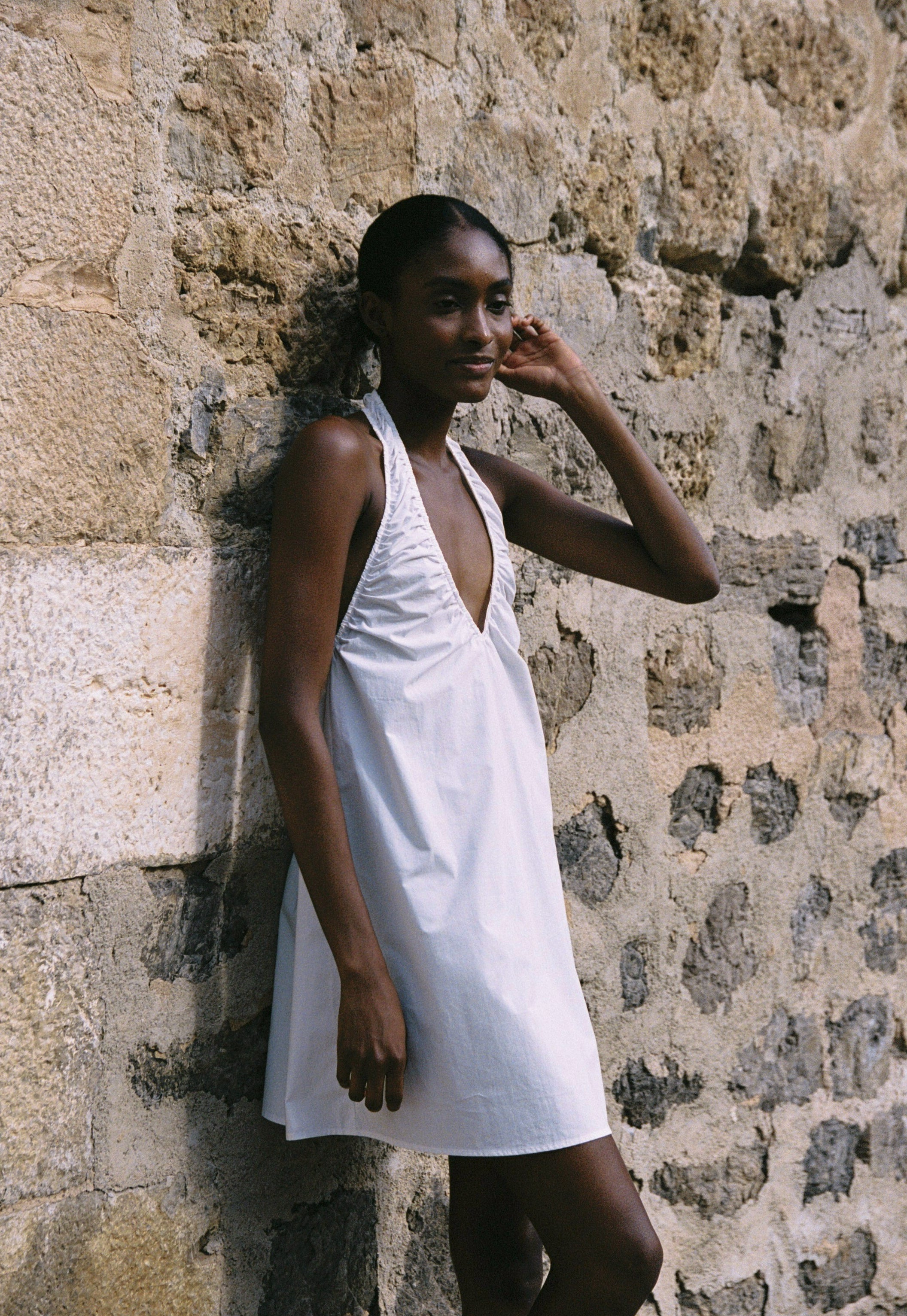 Model wearing the AUDĒRE. Azura cotton halter mini dress in White, side view beside a stone wall.