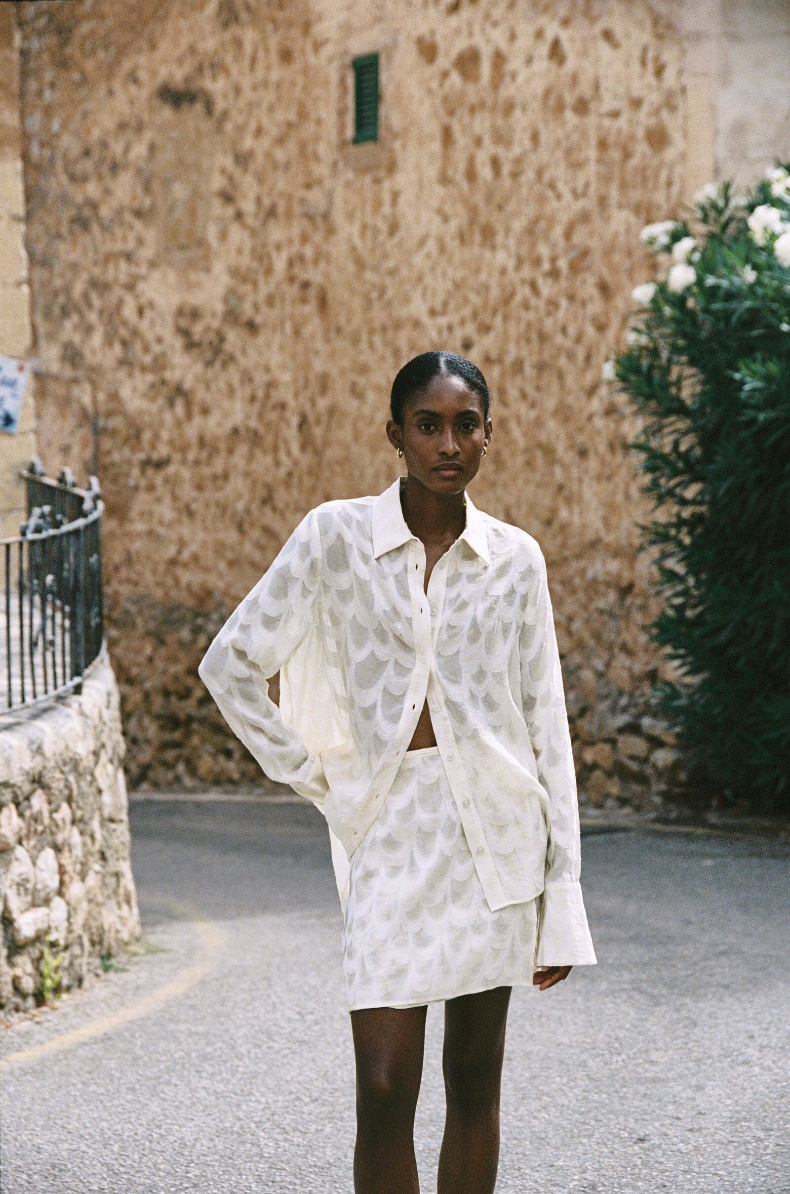 Model wearing the AUDĒRE. Rae semi-sheer shirt and wrap mini skirt in Ivory, standing beside a flowering wall.
