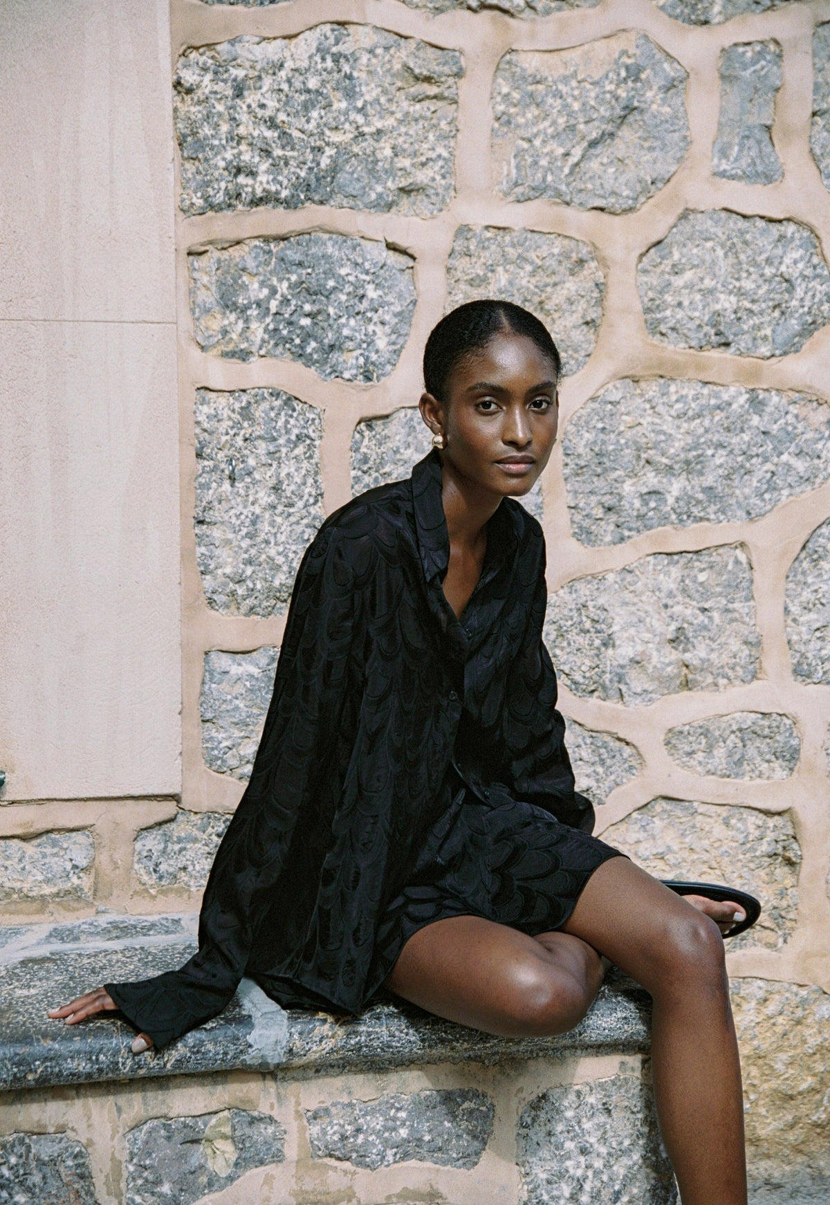 Model wearing the AUDĒRE. Rae semi-sheer shirt in Black, seated on stone steps.