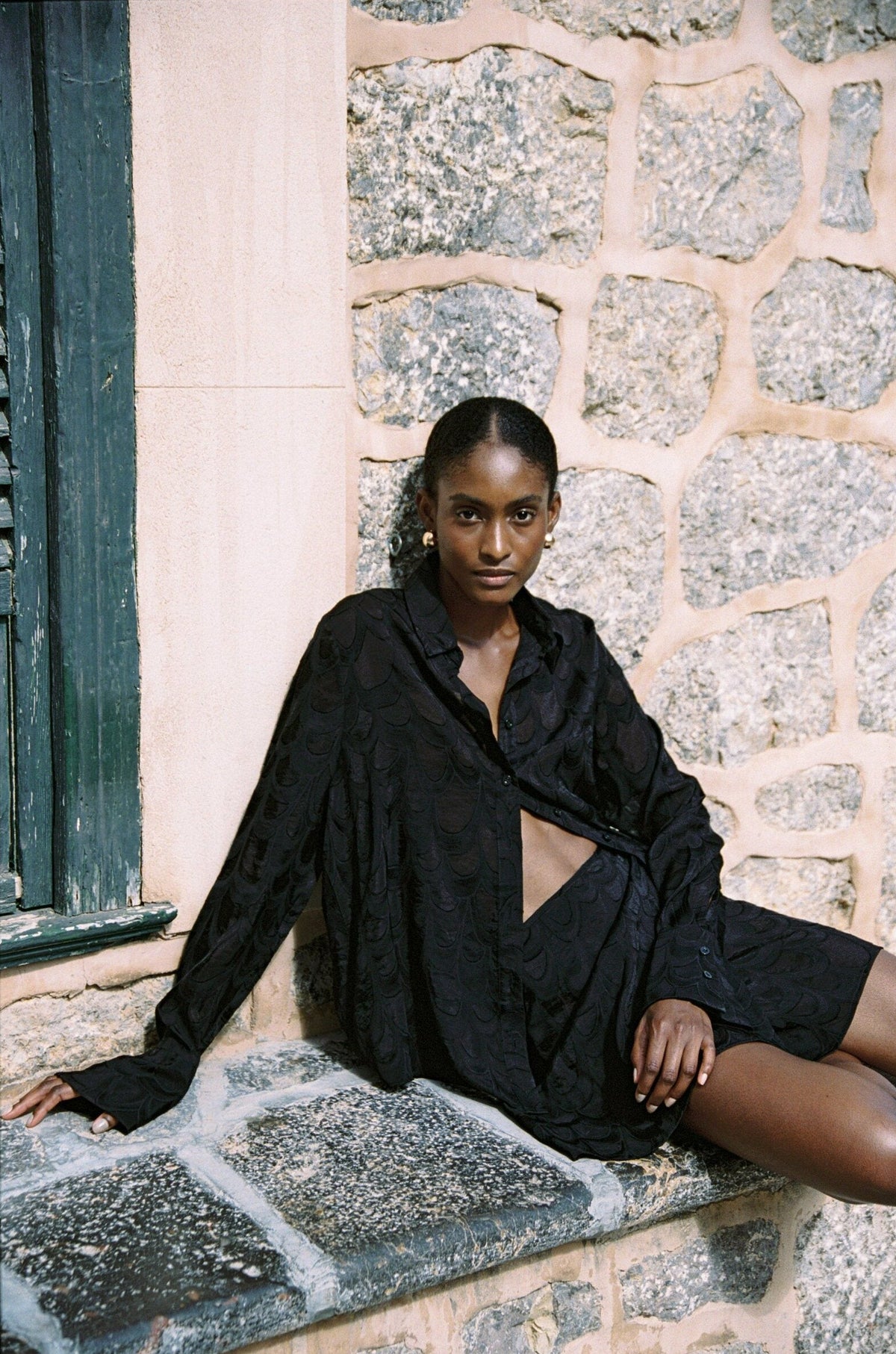 Model wearing the AUDĒRE. Rae semi-sheer shirt in Black, seated with one leg extended on stone steps.