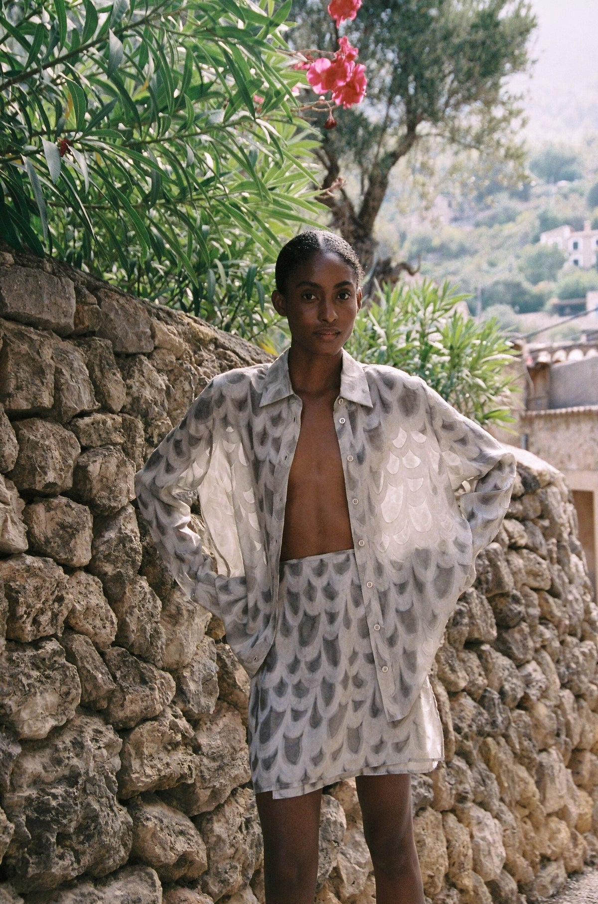 Model wearing the AUDĒRE. Rae semi-sheer shirt in Dove, standing beside a stone wall with the matching wrap mini skirt.