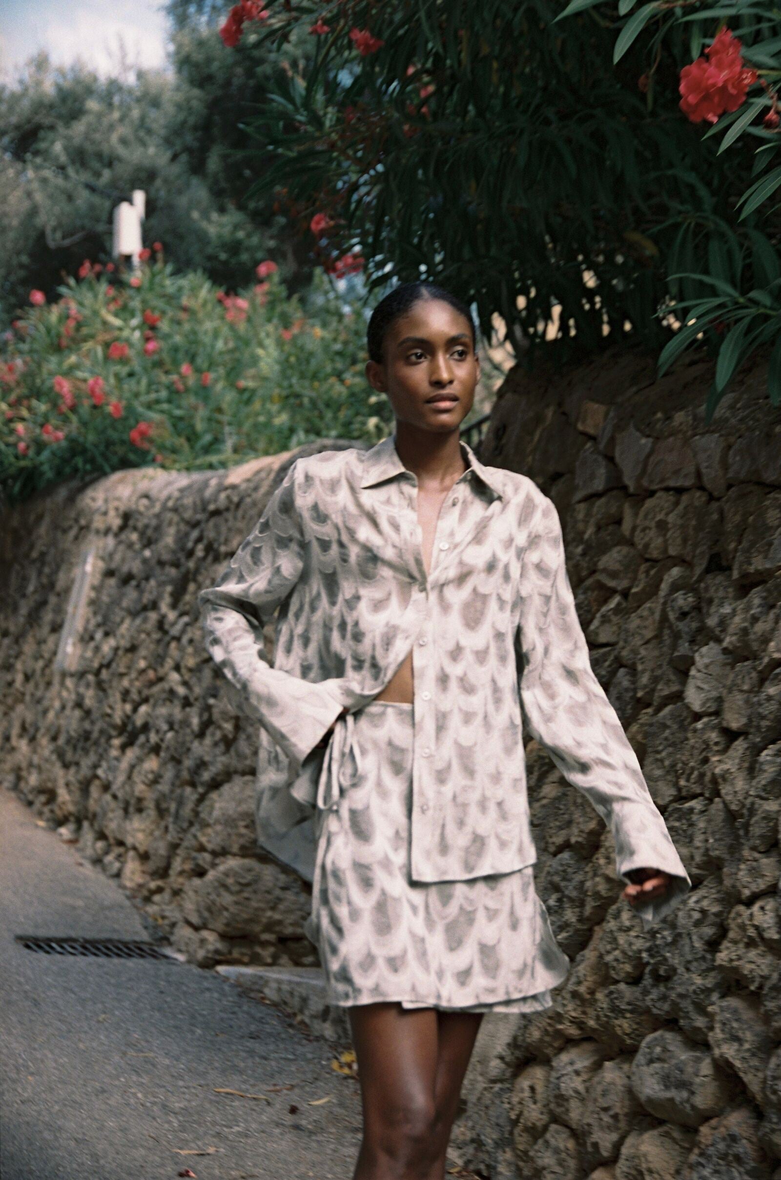 Model wearing the AUDĒRE. Rae semi-sheer shirt in Dove, standing beside a flowering wall.