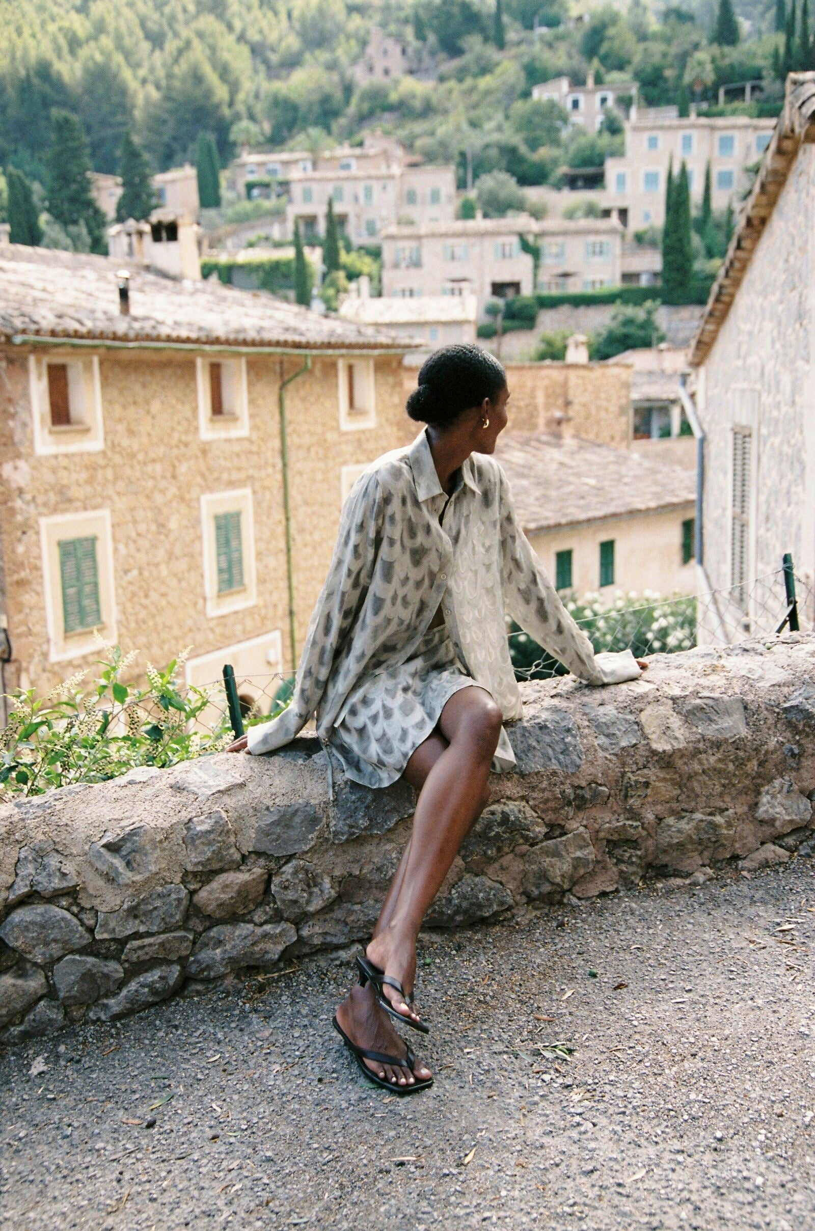 Model wearing the AUDĒRE. Rae semi-sheer shirt in Dove, full-length view seated on a stone wall.