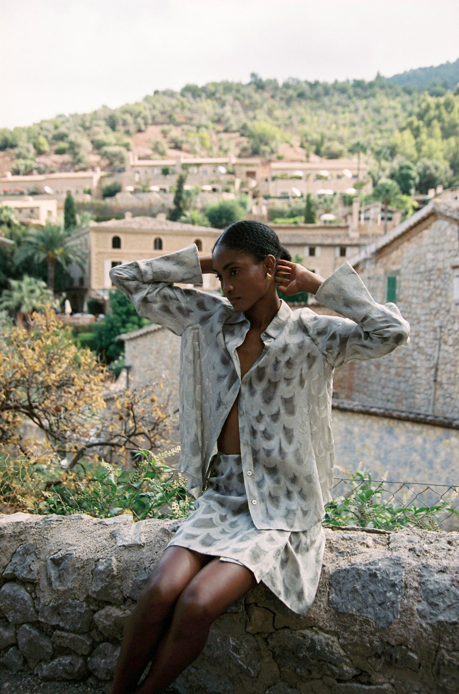 Model wearing the AUDĒRE. Rae semi-sheer shirt in Dove, seated with arms raised overlooking a village.