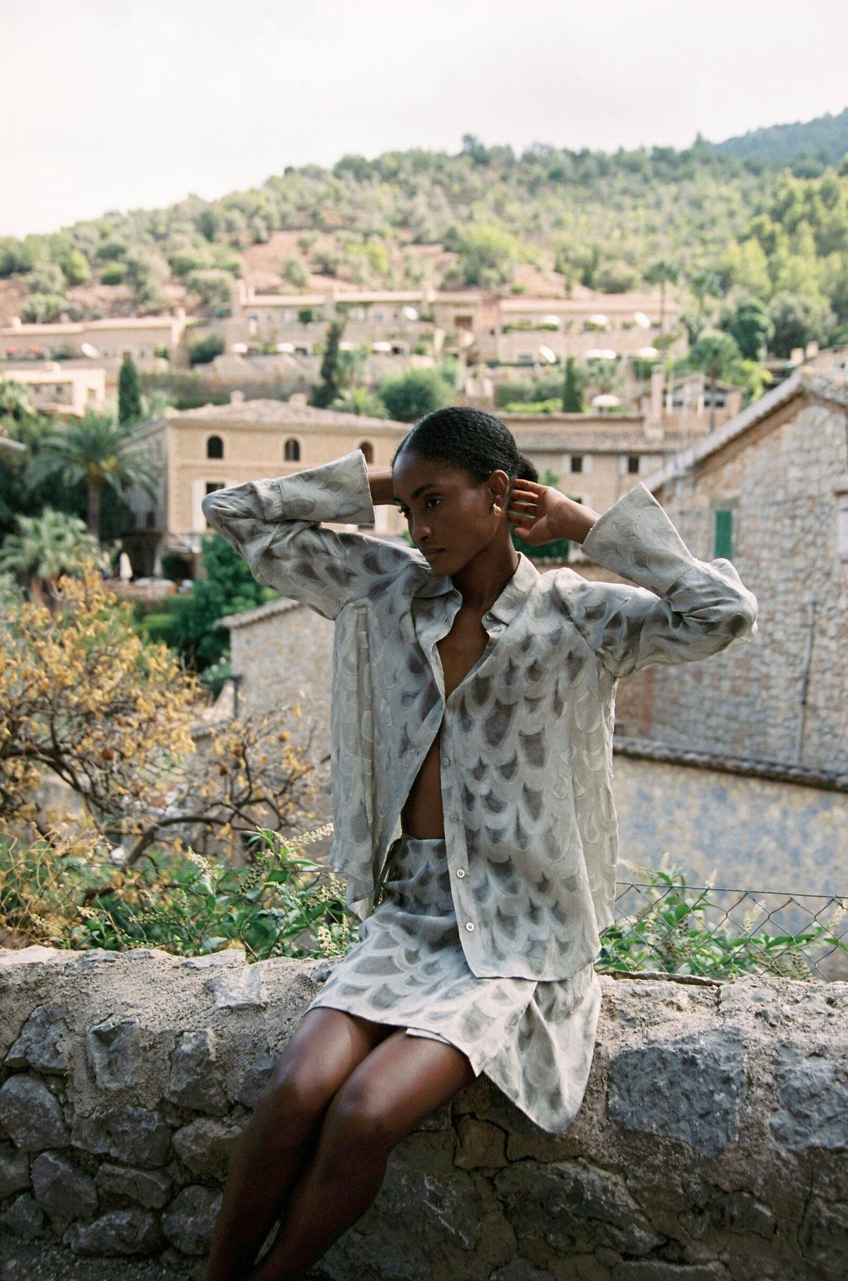 Model wearing the AUDĒRE. Rae semi-sheer shirt in Dove, seated with arms raised overlooking a village.
