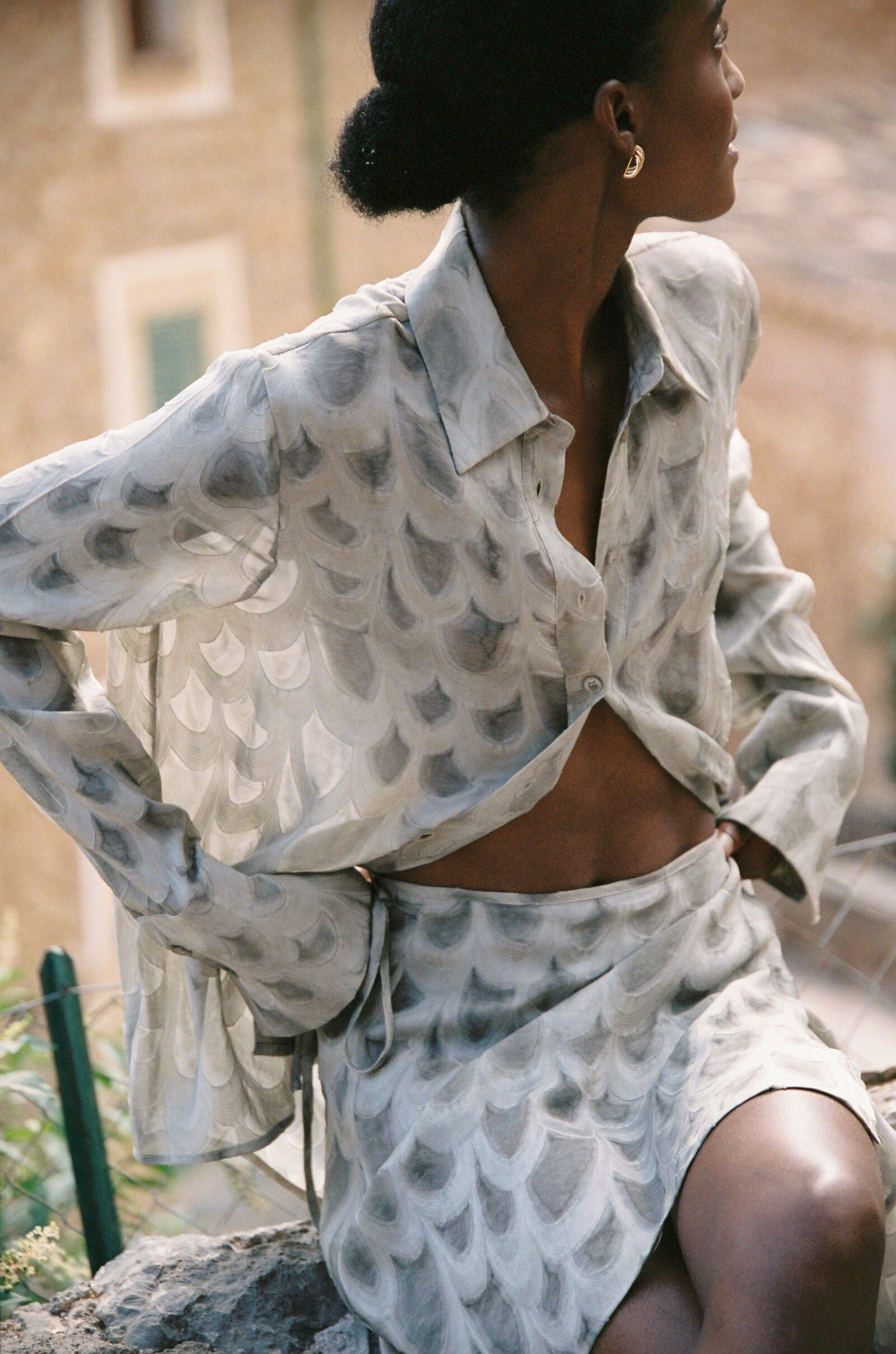 Model wearing the AUDĒRE. Rae semi-sheer shirt in Dove, side view seated on a stone wall.