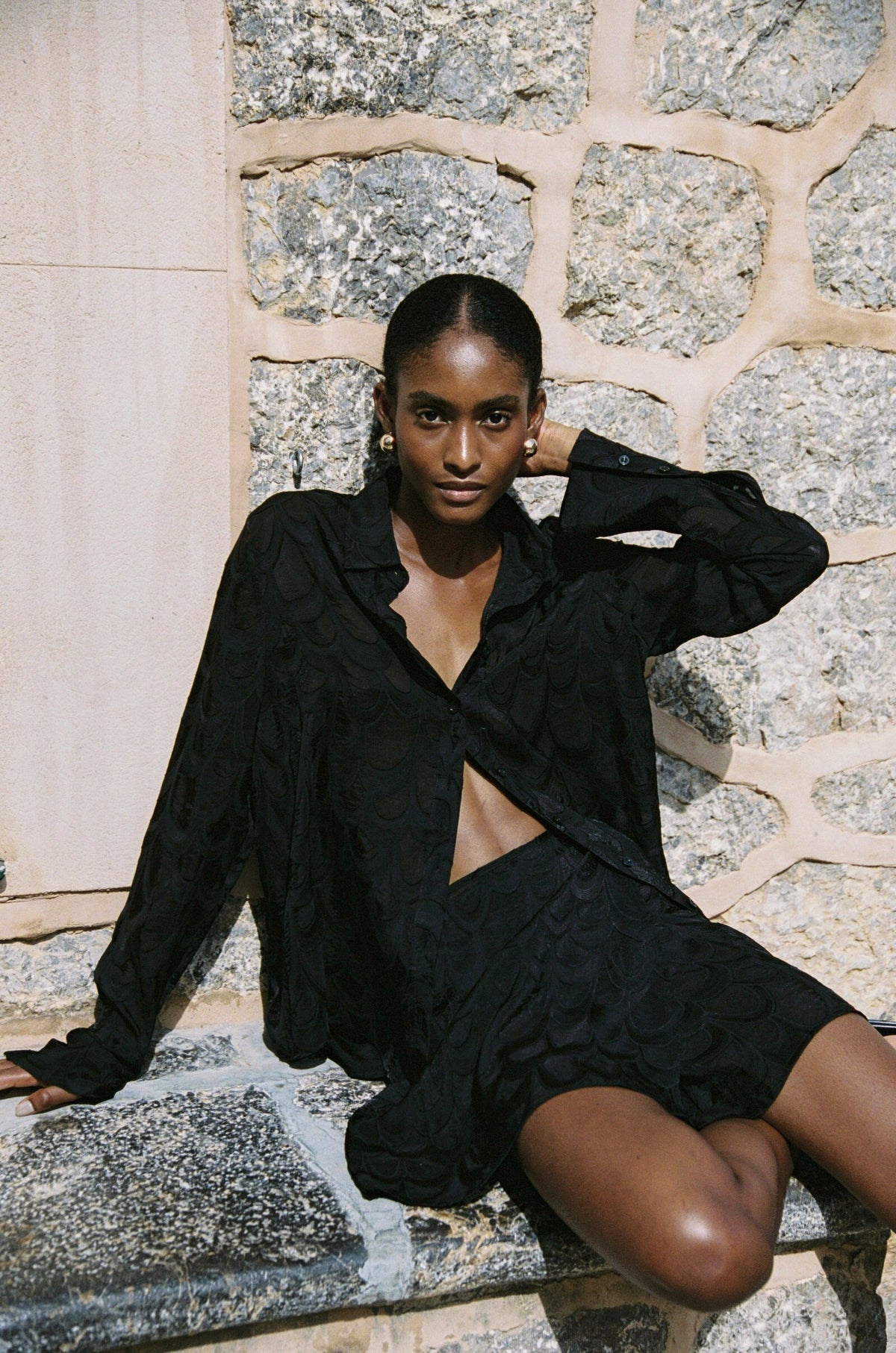 Model wearing the AUDĒRE. Rae wrap mini skirt in Black, seated on stone steps with the matching shirt.