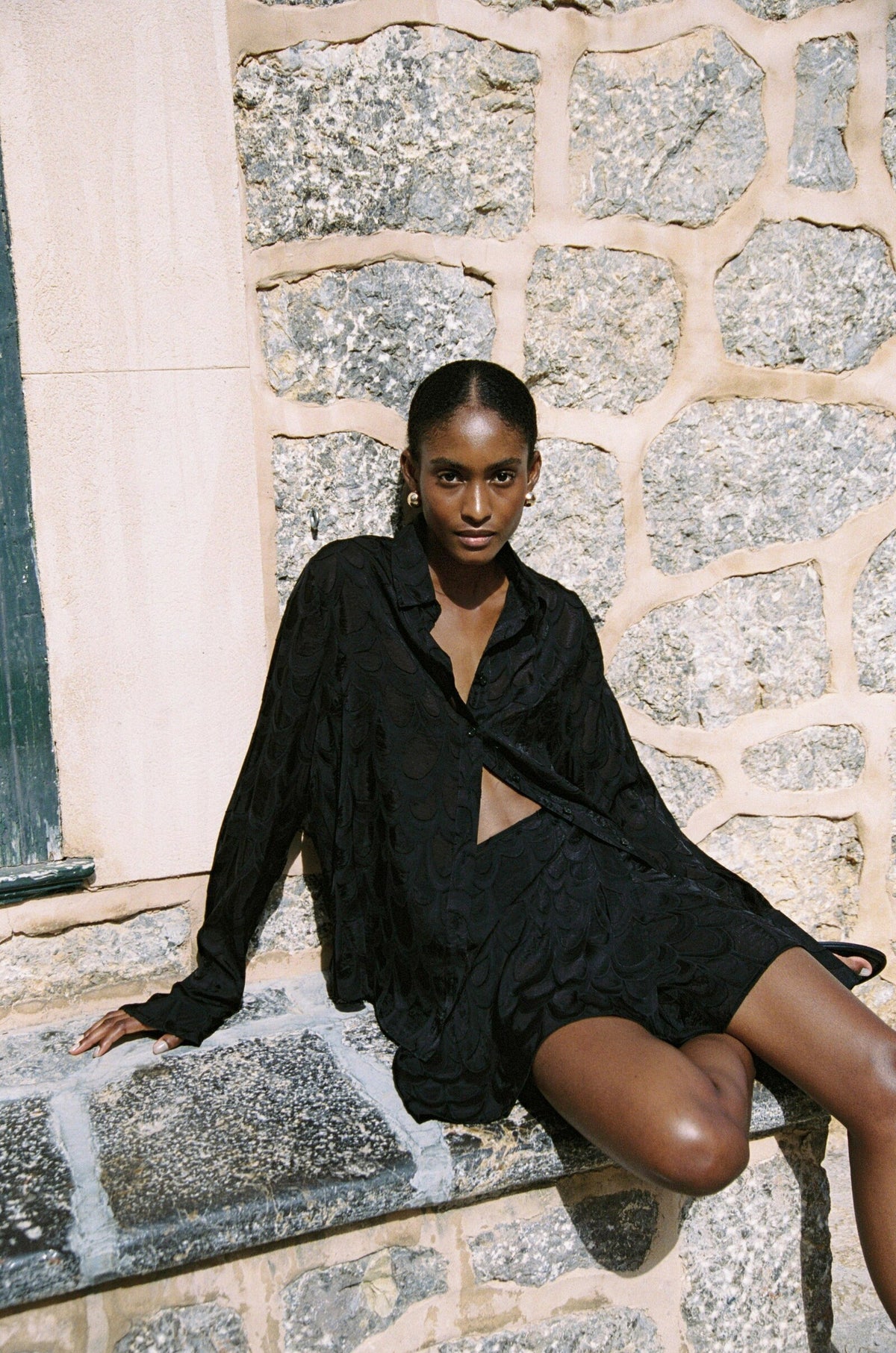 Model wearing the AUDĒRE. Rae wrap mini skirt in Black, seated on stone steps.