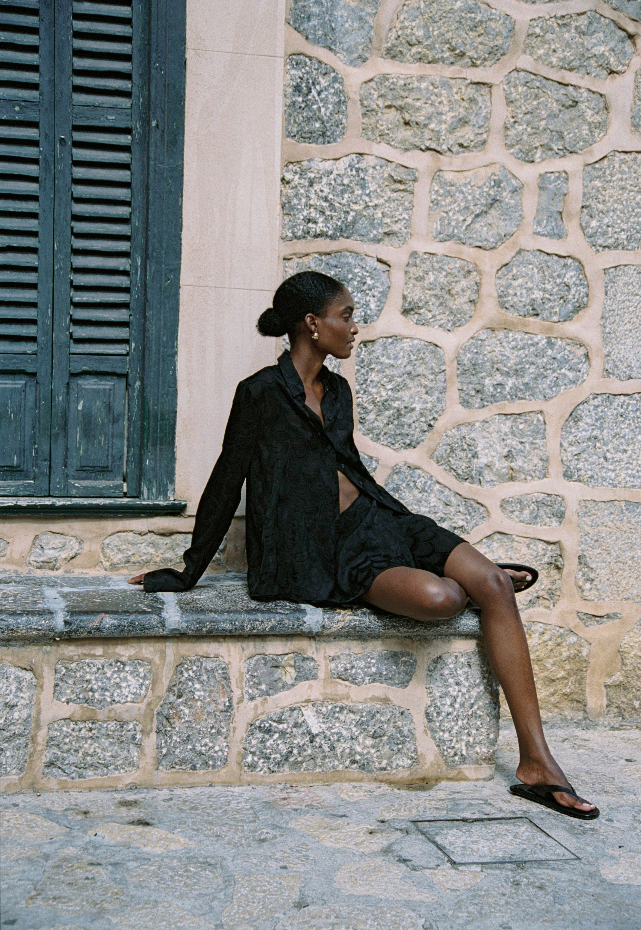 Model wearing the AUDĒRE. Rae wrap mini skirt in Black, seated beside green shutters.