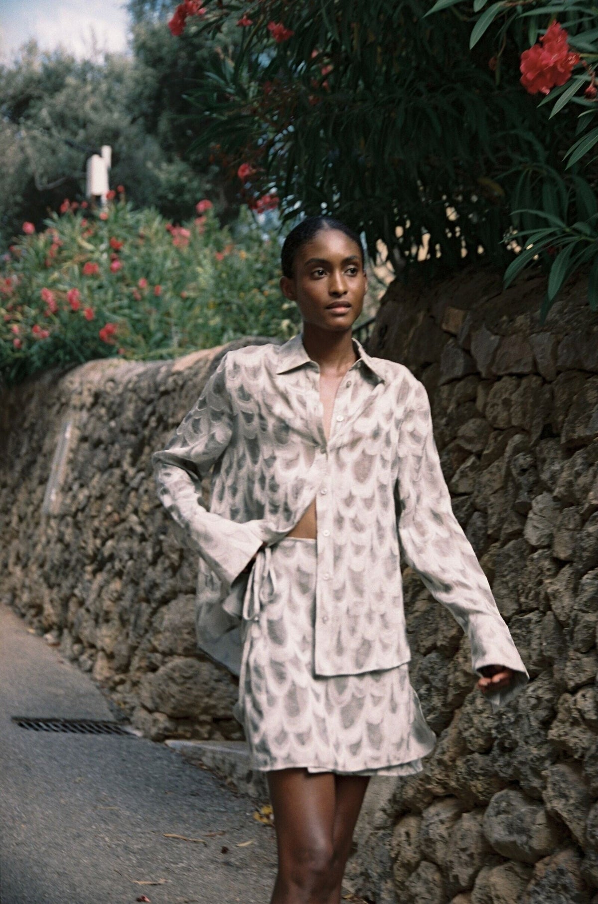 Model wearing the AUDĒRE. Rae wrap mini skirt in Dove, standing beside a flowering stone wall.