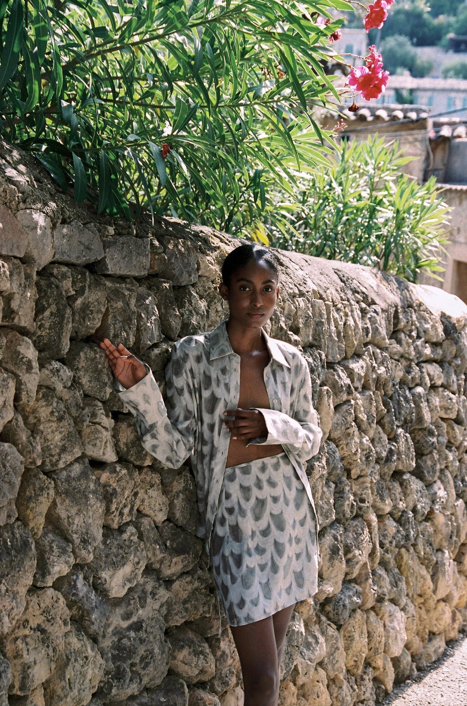 Model wearing the AUDĒRE. Rae wrap mini skirt in Dove, standing beside a stone wall with the matching shirt.