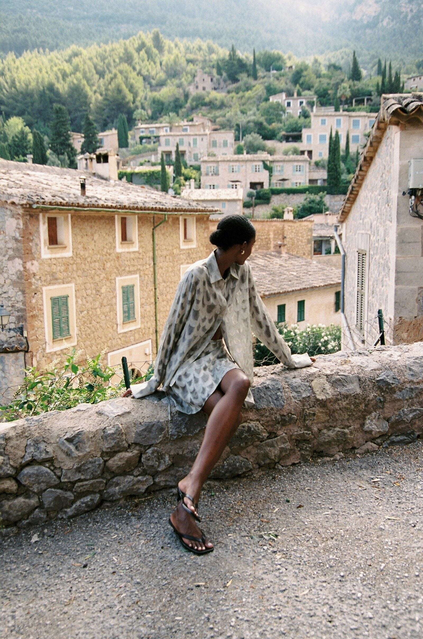 Model wearing the AUDĒRE. Rae wrap mini skirt in Dove, seated on a stone wall overlooking a village.