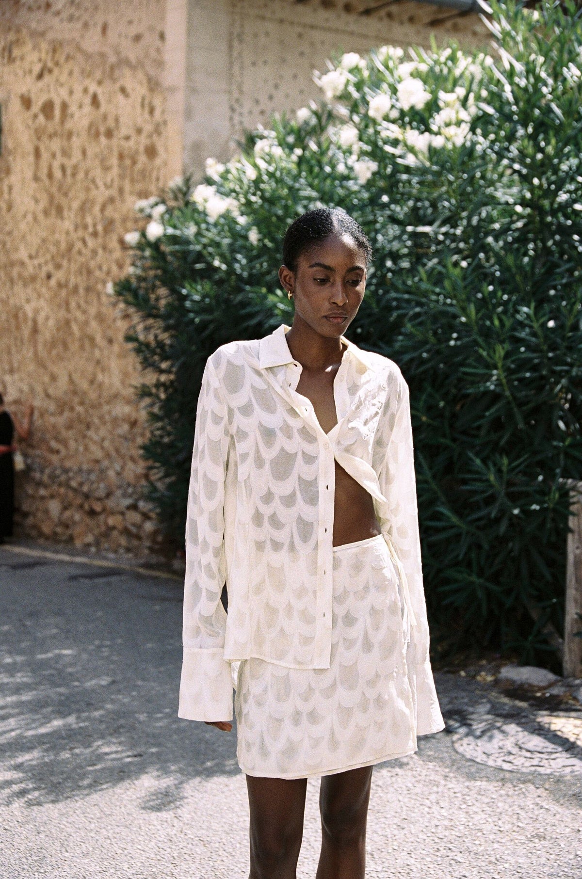 Model wearing the AUDĒRE. Rae wrap mini skirt in Ivory, standing beside a flowering wall with the matching shirt.