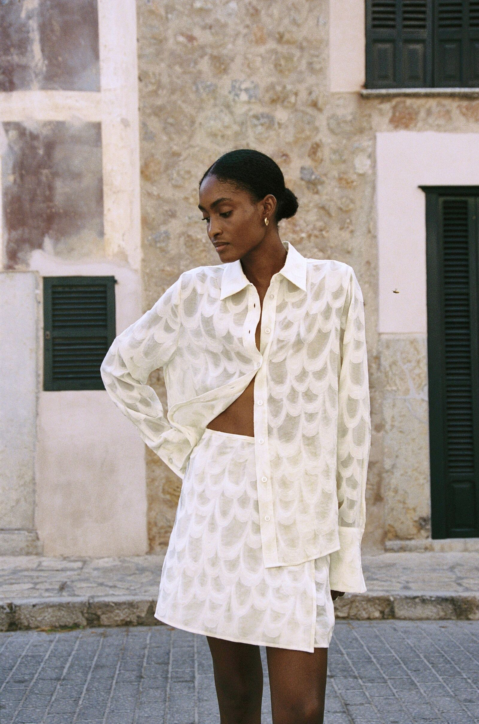 Model wearing the AUDĒRE. Rae wrap mini skirt in Ivory, standing beside green shutters.