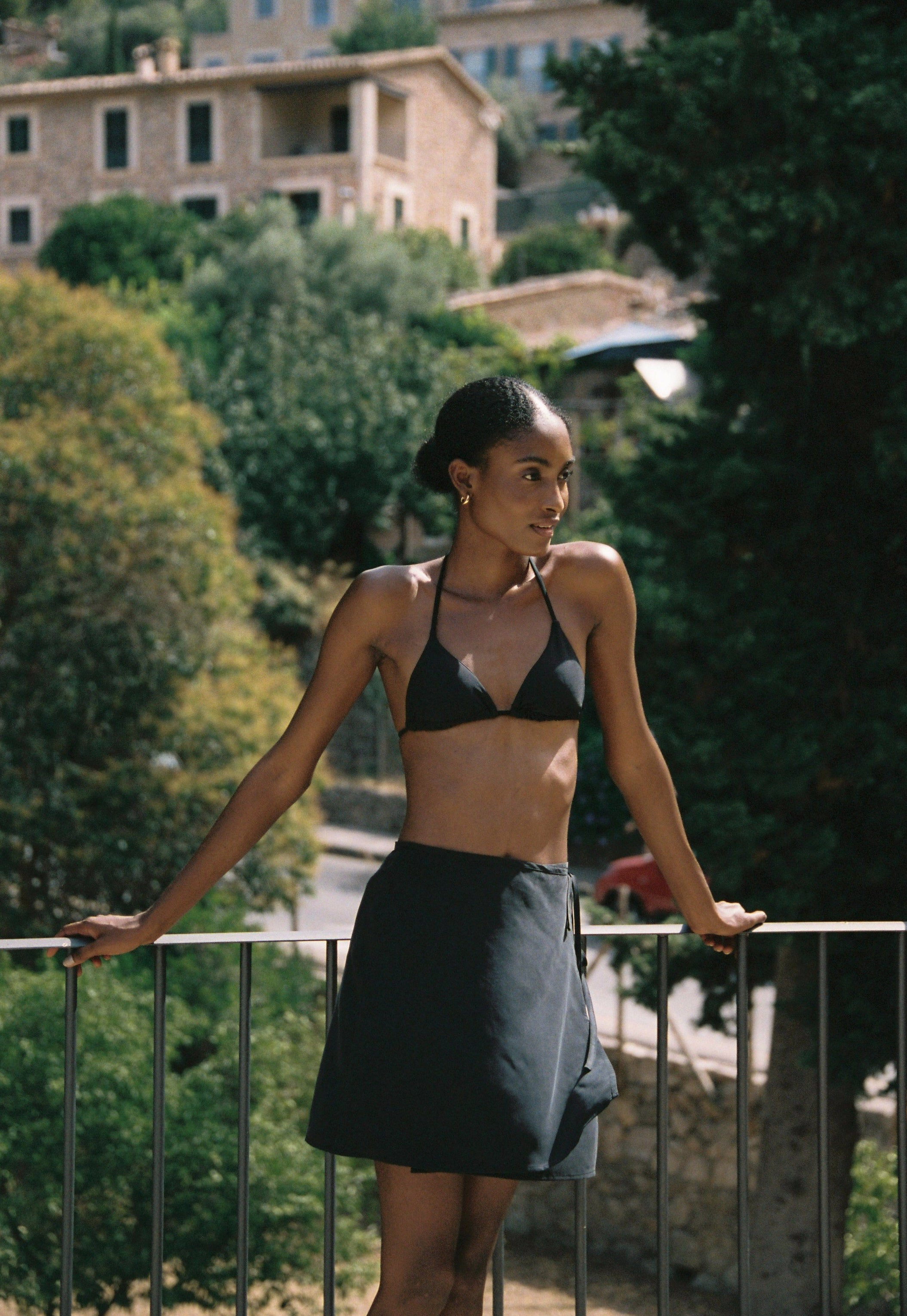 Model wearing the AUDĒRE. Vero cupro-blend wrap mini skirt in Black, front view beside a railing.