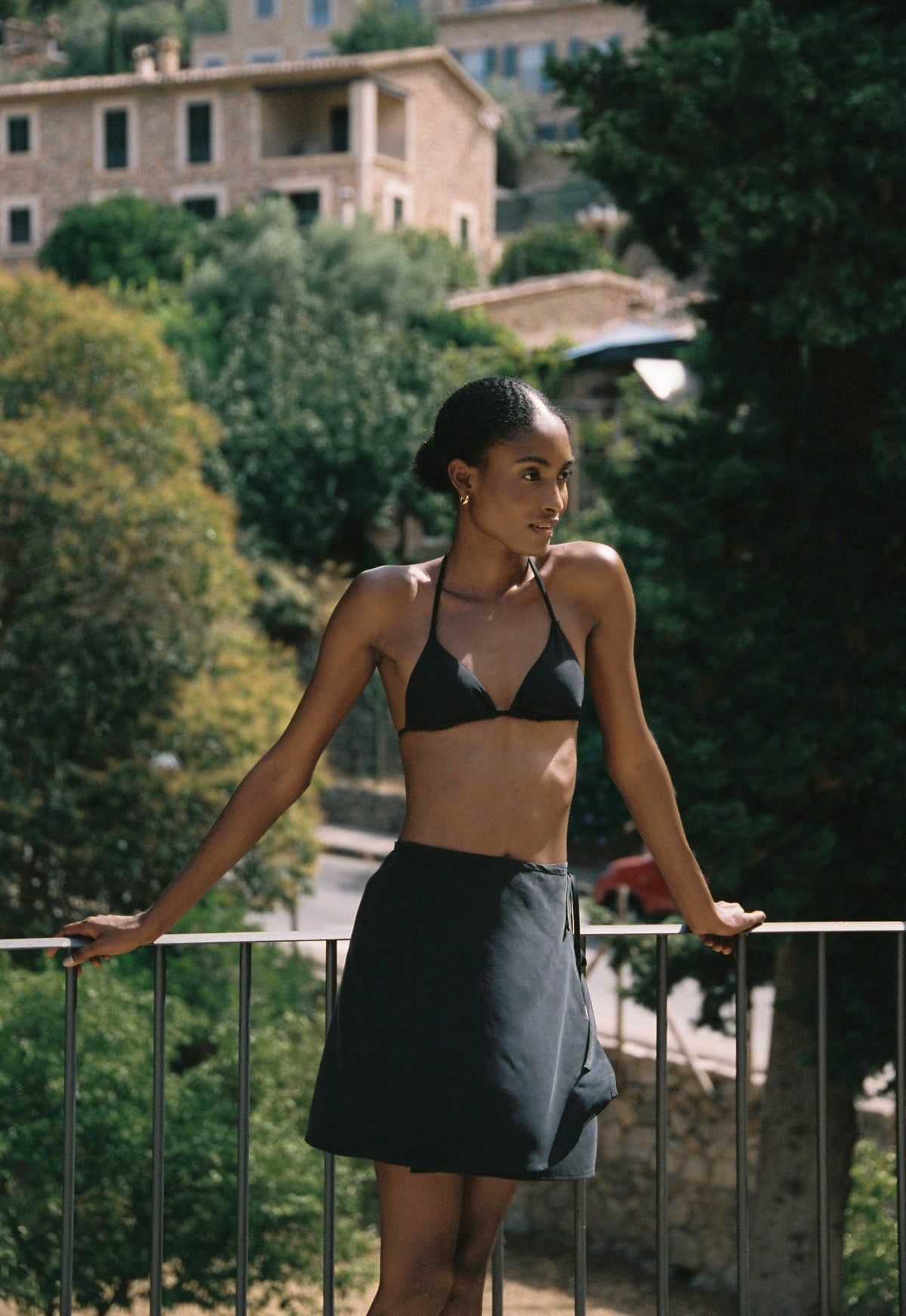 Model wearing the AUDĒRE. Vero cupro-blend wrap mini skirt in Black, front view beside a railing.