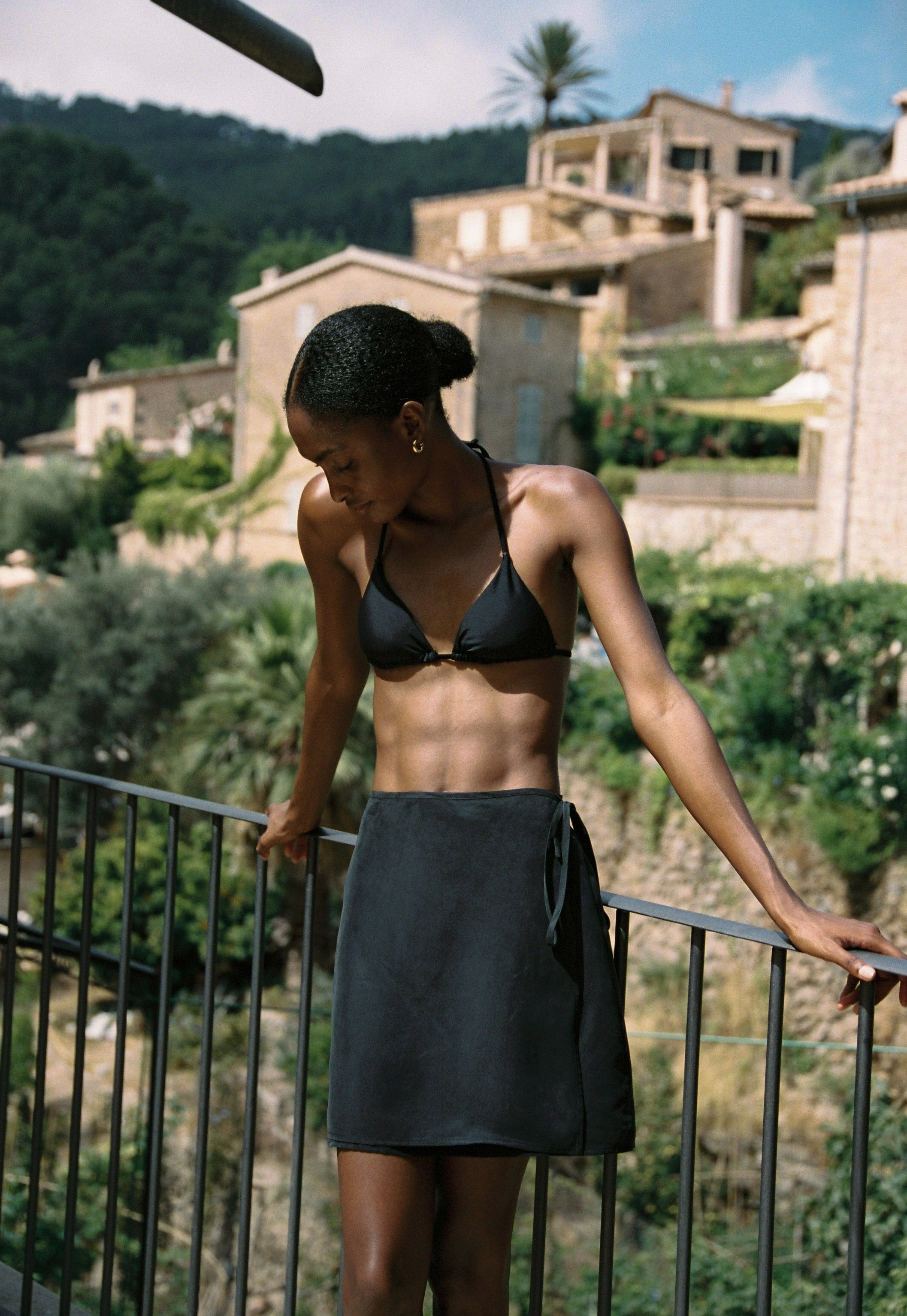 Model wearing the AUDĒRE. Vero cupro-blend wrap mini skirt in Black, side view beside a railing.